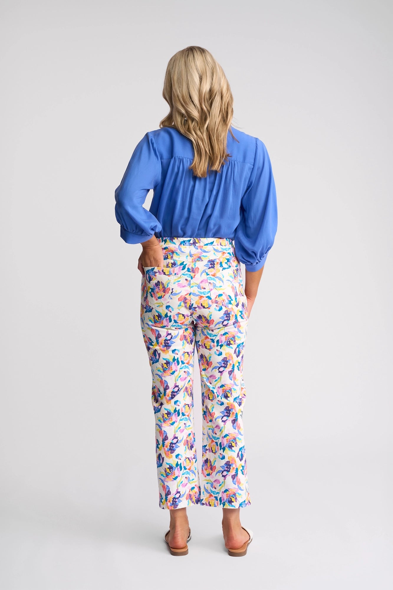 A woman with blonde hair stands facing away, wearing a blue blouse, Vassalli Seattle Pull On - Demi stretch cotton floral print pants, and sandals against a plain light background.