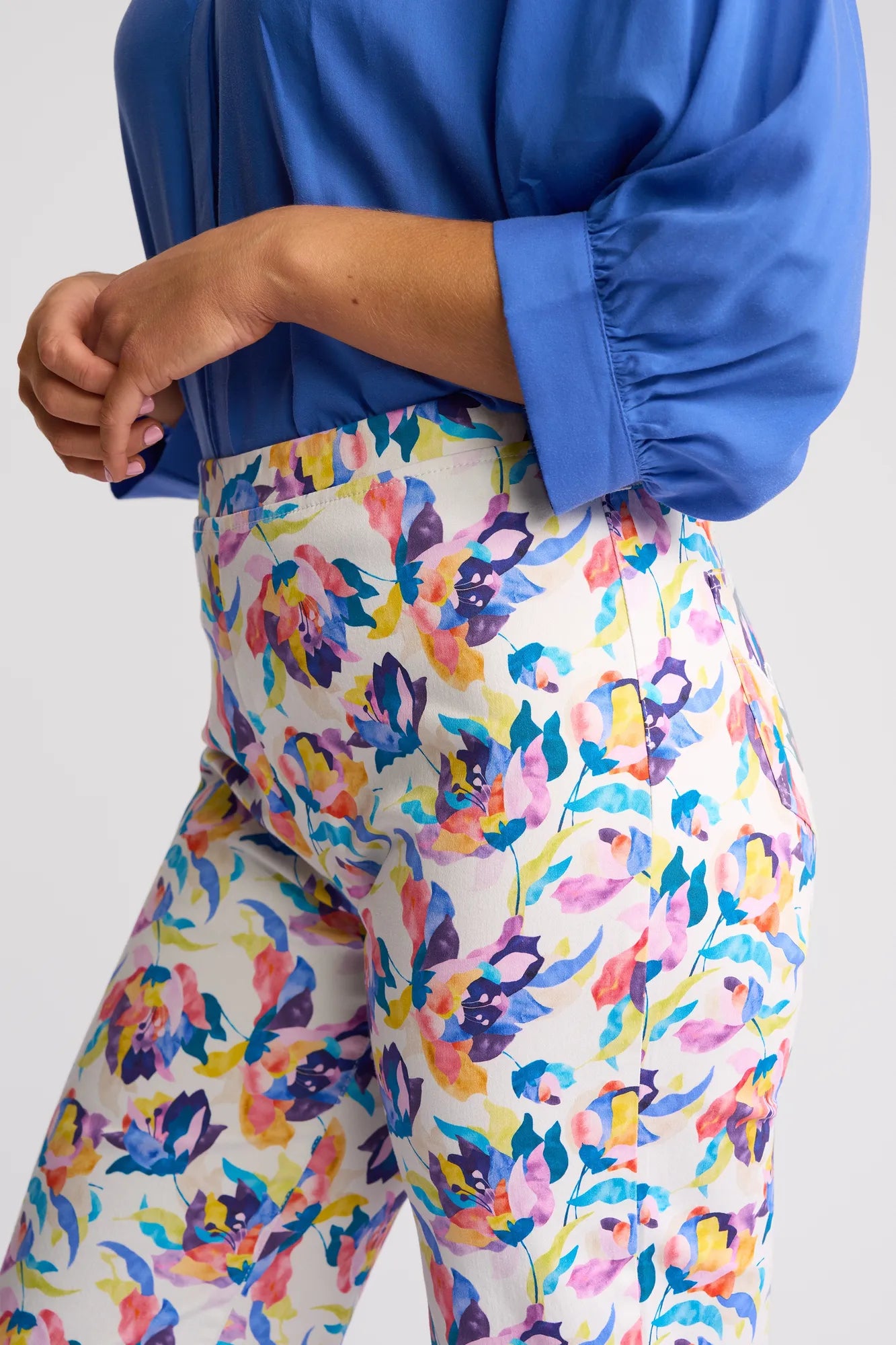 A person wears a blue blouse with the sleeves rolled up and Vassalli’s Seattle Pull On - Demi high-waisted pants, which feature a colorful, abstract floral print in pink, purple, yellow, and blue on a white background.