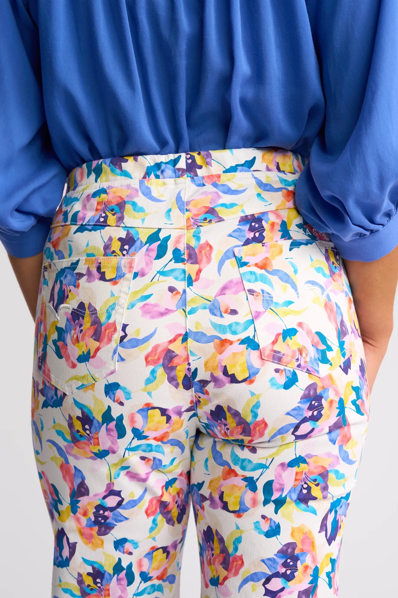 A person is shown from the back wearing a blue shirt and Vassalli’s Seattle Pull On - Demi pants, which feature a white background with colorful purple, yellow, pink, and blue floral patterns, visible back pockets, and an easy pull-on design.