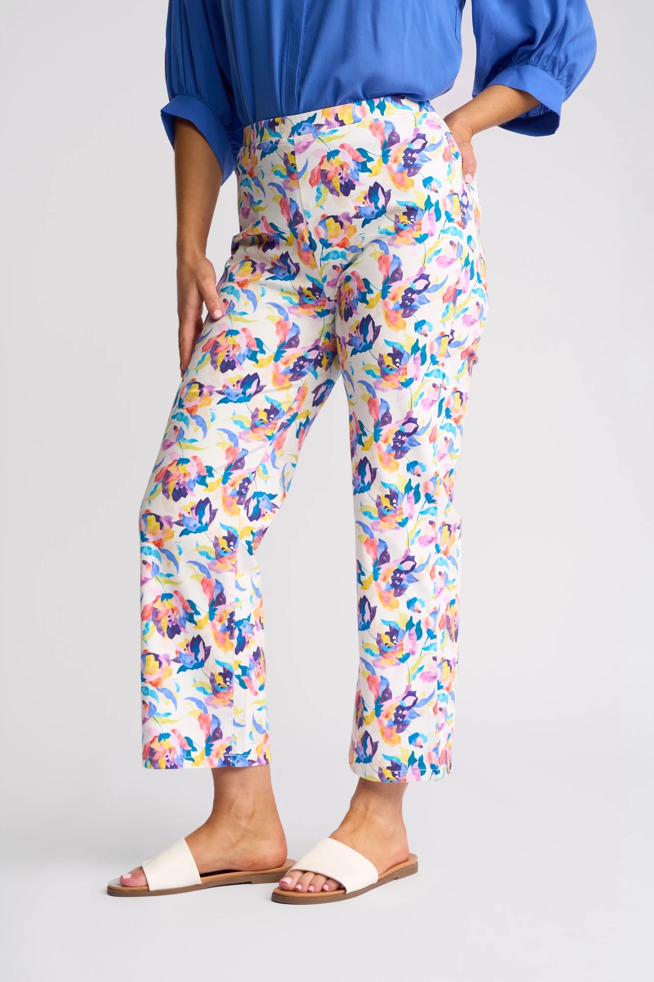 A person wearing the Vassalli Seattle Pull On - Demi pants in a colorful floral print, paired with a blue shirt with rolled sleeves and white sandals, stands against a plain light background.