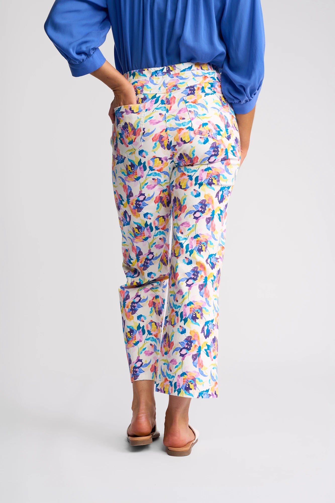 A person wearing a blue top, the Vassalli Seattle Pull On - Demi colorful floral print pants, and brown sandals stands with one hand in their back pocket, facing away from the camera against a plain background.