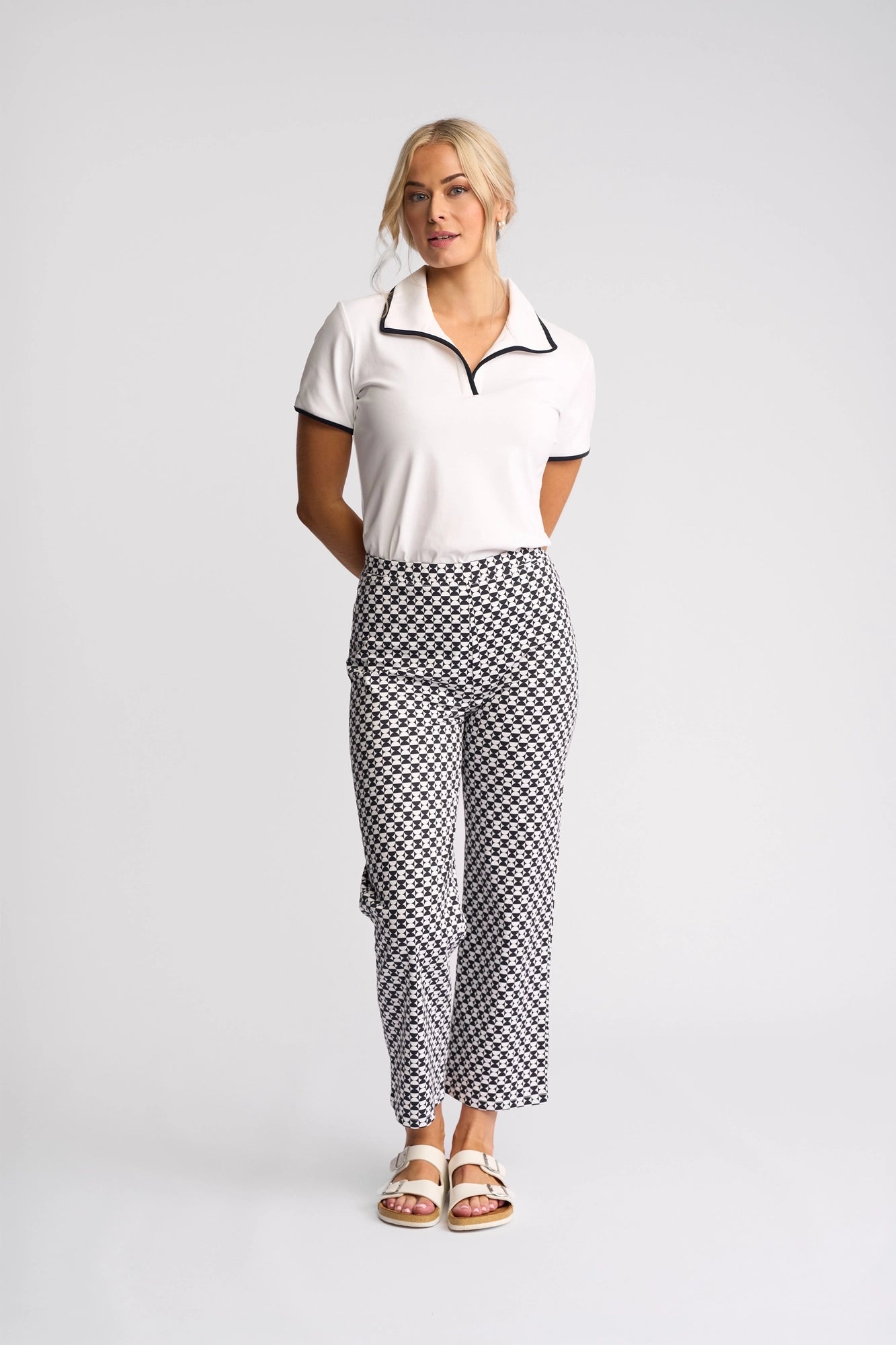A woman stands against a plain background, wearing a white collared shirt with black trim, Vassalli Seattle Pull On - Dixie stretch cotton pants featuring a geometric pattern, and white sandals. Her hands are behind her back, and she is looking slightly to the side.