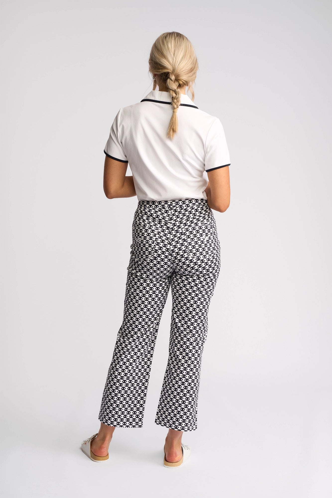 A woman with blonde braided hair, dressed in a white collared shirt and Vassalli Seattle Pull On - Dixie stretch cotton pants featuring a black-and-white geometric pattern, stands facing away against a plain white background.