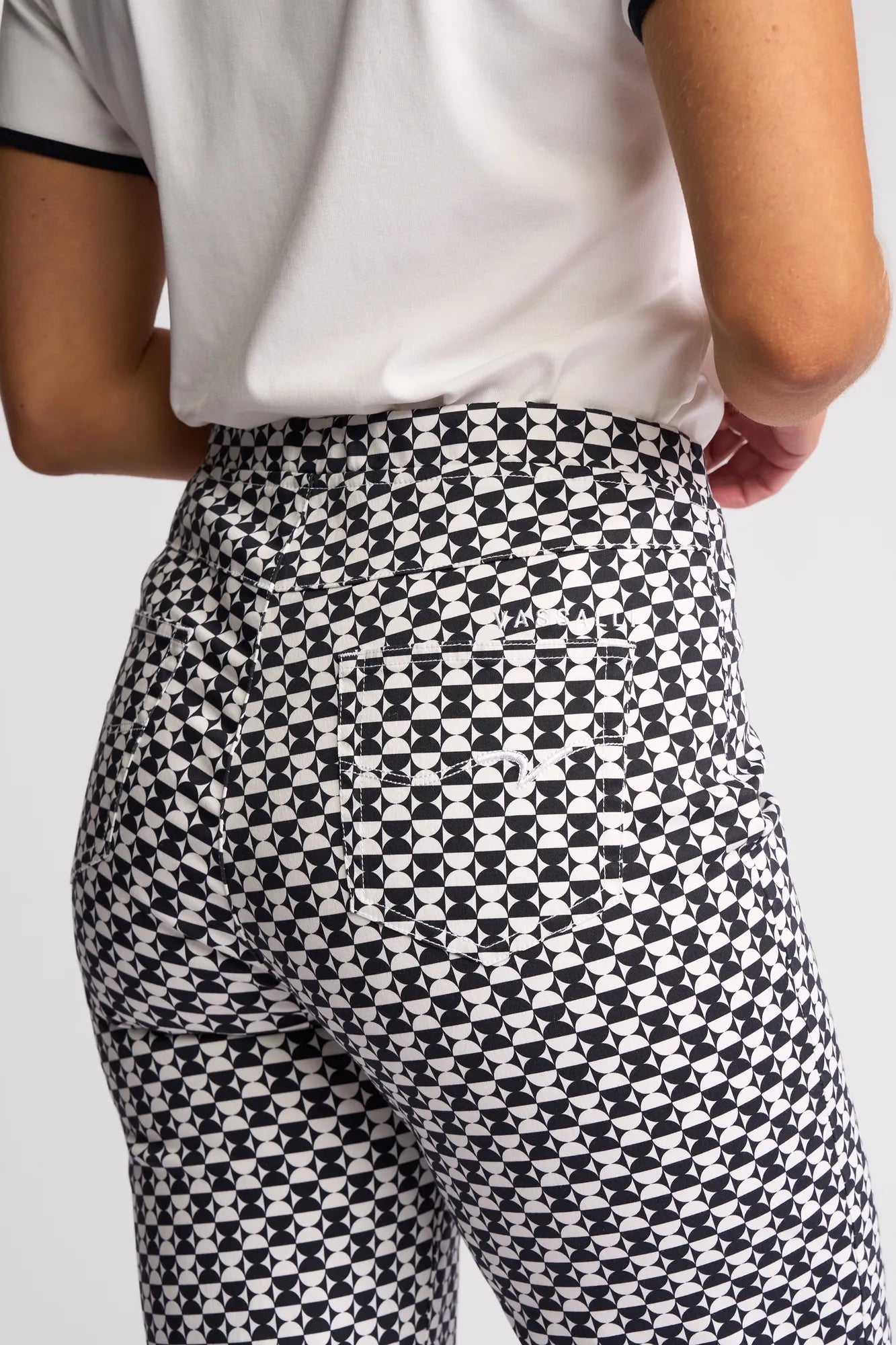 A person is seen from the back and waist up wearing Vassalli’s Seattle Pull On - Dixie pants, featuring a bold black and white geometric pattern crafted from stretch cotton for comfort, paired with a white short-sleeve shirt that highlights the striking design of these pants.
