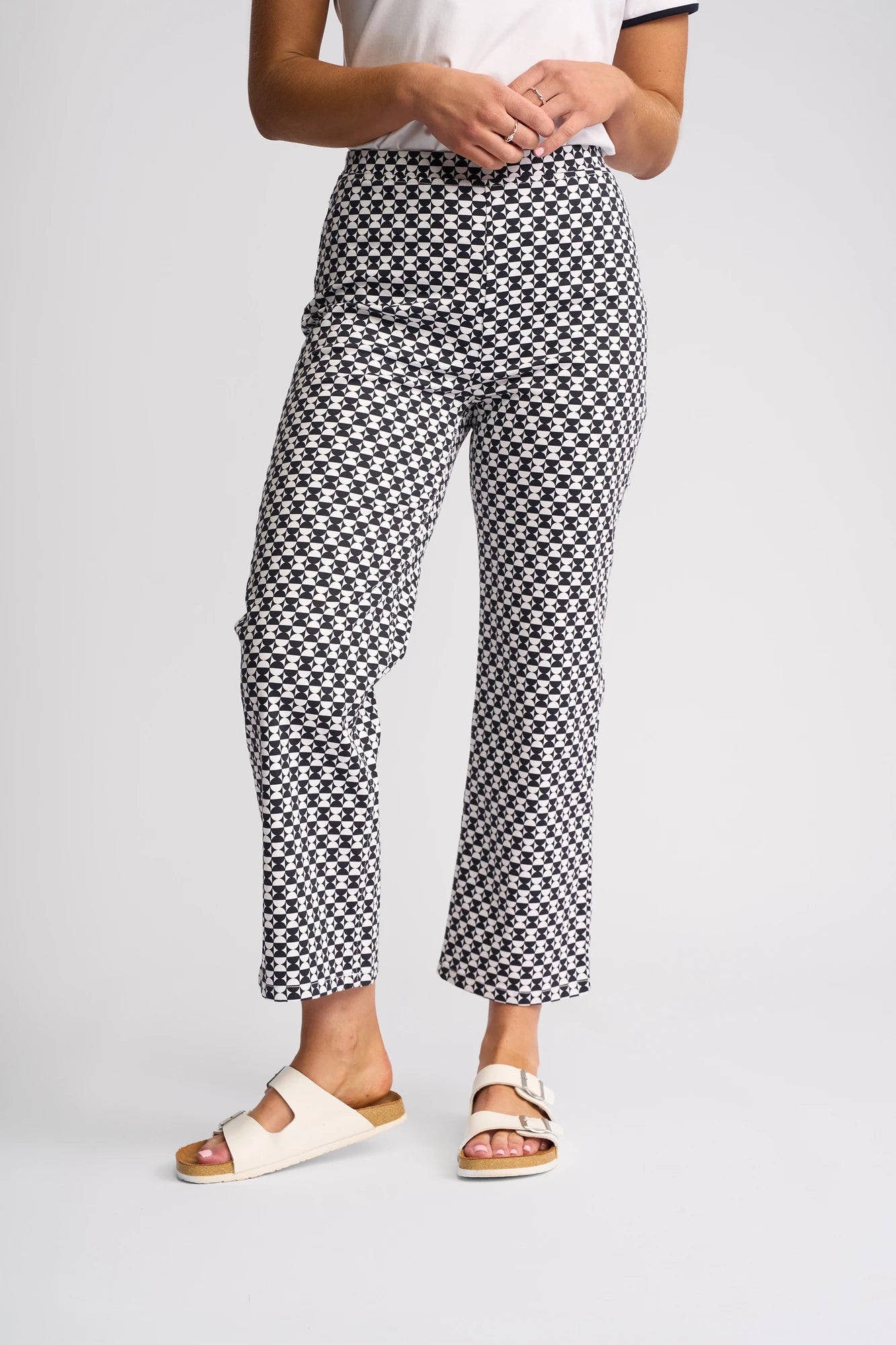 A person stands against a plain white background wearing Vassalli Seattle Pull On - Dixie pants with a black and white geometric checkered pattern, paired with a white short-sleeve top and white sandals. Their upper face is not visible.