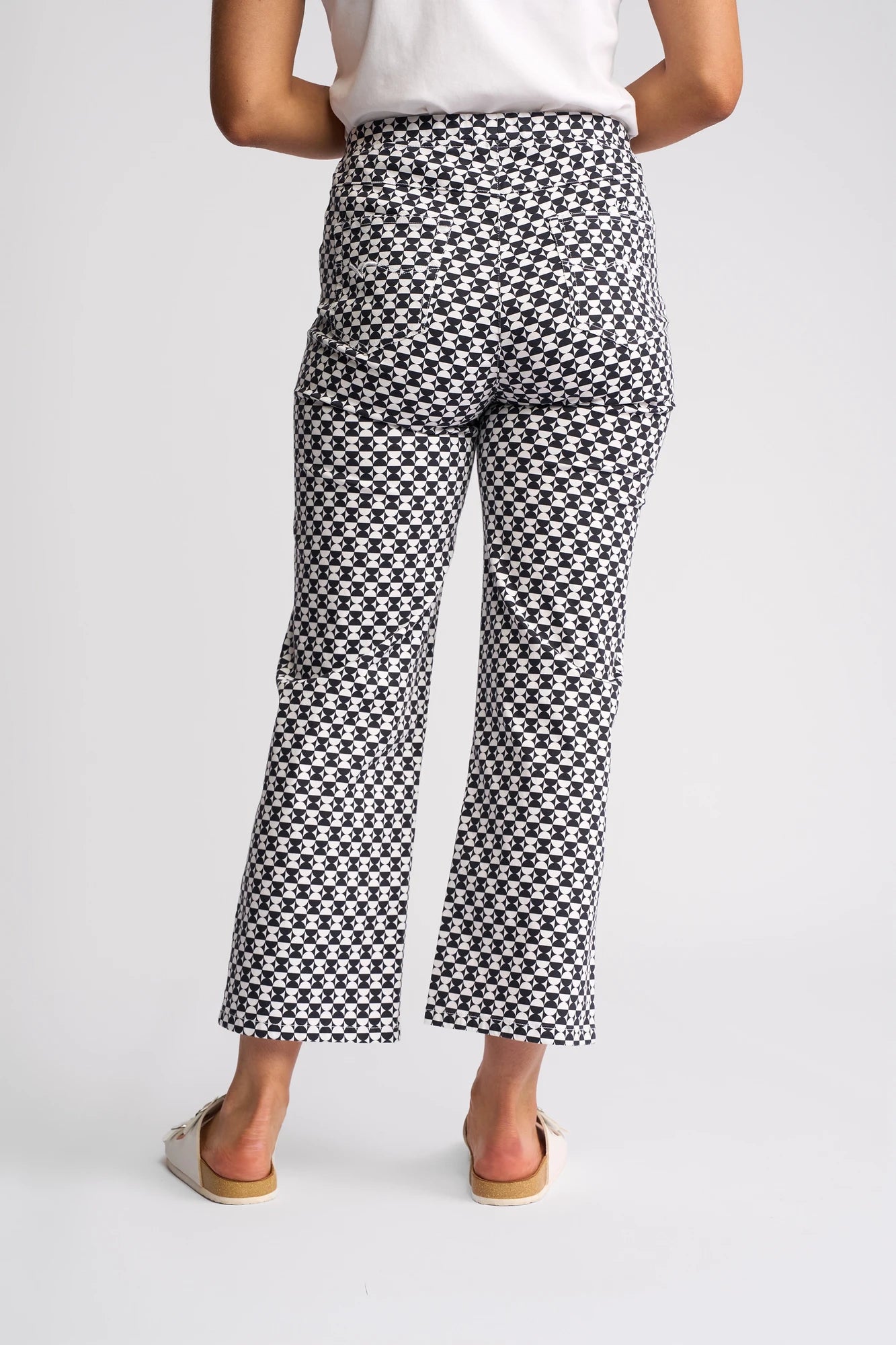 A person is shown from behind against a plain light background, wearing Vassalli’s Seattle Pull On - Dixie pants, which feature a bold black and white geometric checkered pattern, paired with white sandals.
