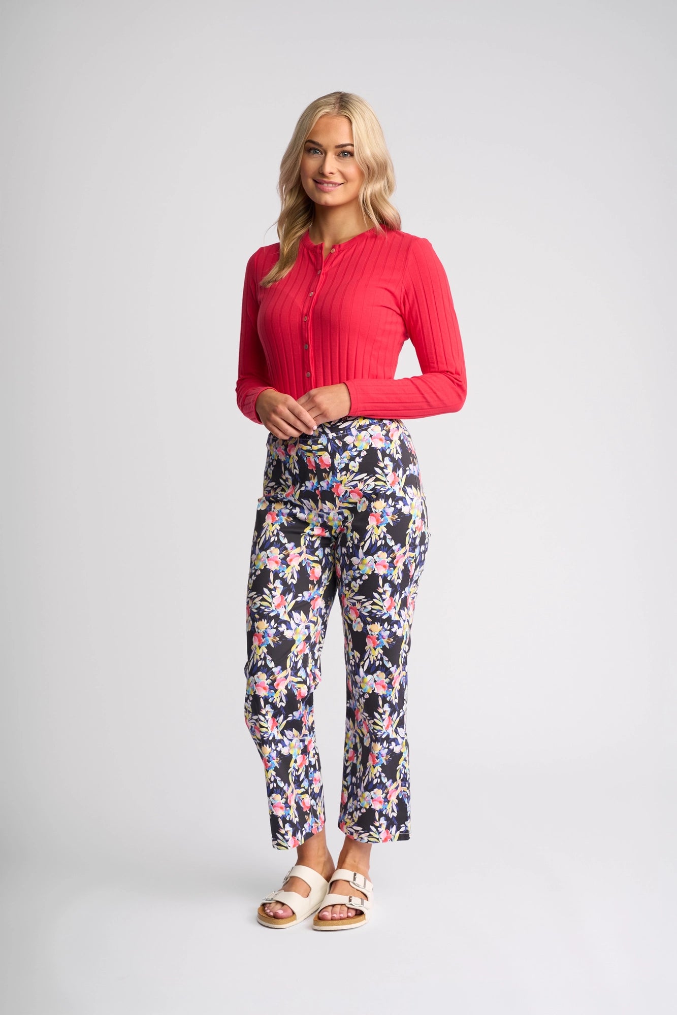 A woman stands smiling against a plain light background, wearing a long-sleeve red top, the Vassalli Seattle Pull On - Lani floral print pant crafted from stretch cotton, and white sandals.