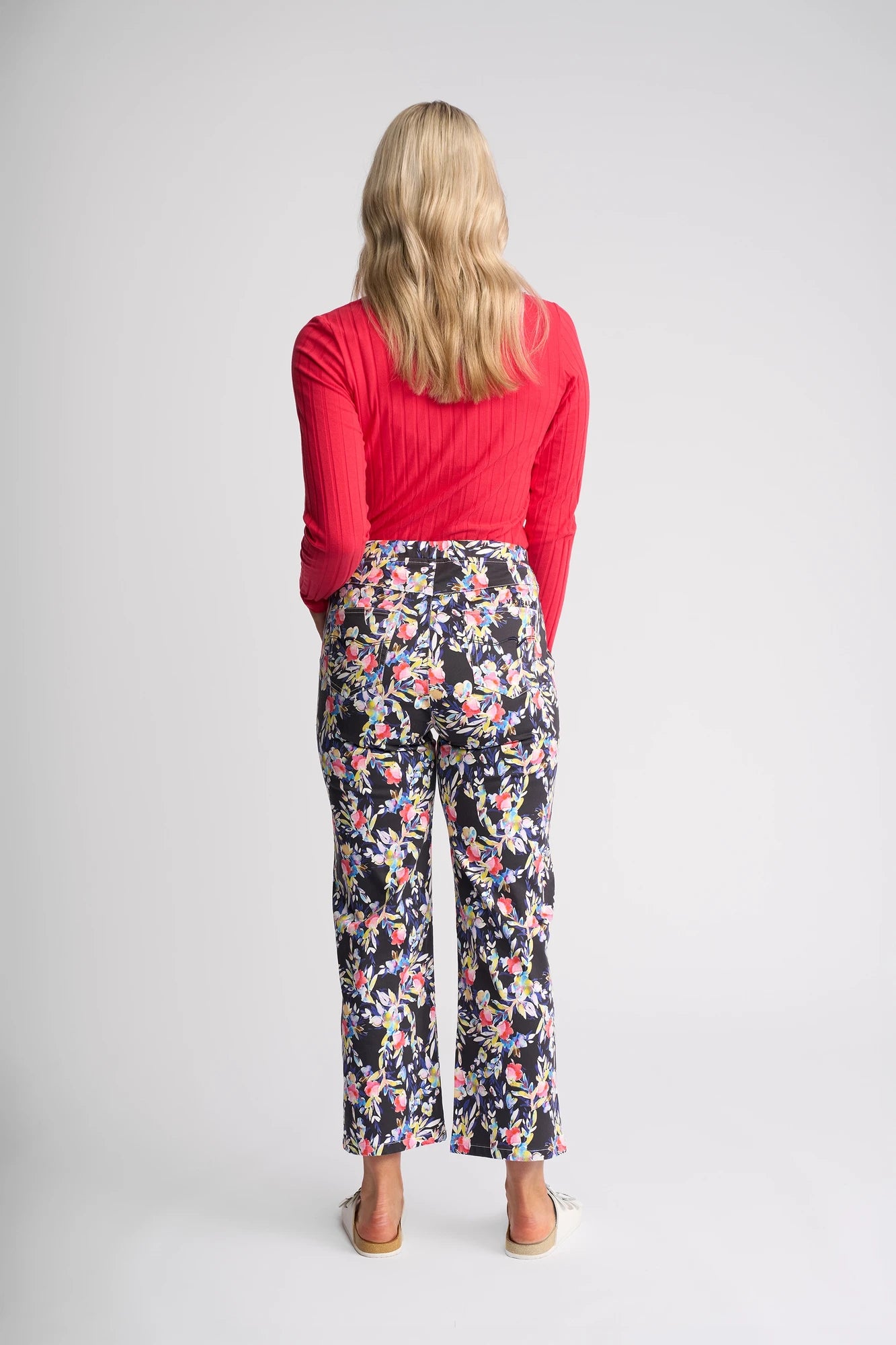 A person with long blonde hair, wearing a red long-sleeve top and Vassalli’s Seattle Pull On - Lani floral print stretch cotton pants, stands facing away from the camera against a plain light background.