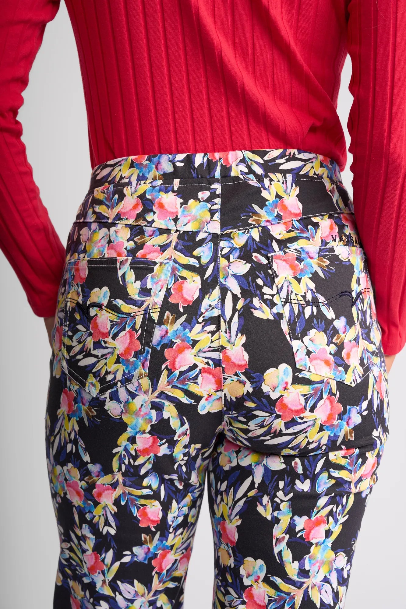 A person wearing a red long-sleeve top and the Vassalli Seattle Pull On - Lani high-waisted, black floral print pant made from stretch cotton, shown from the back in a relaxed style.