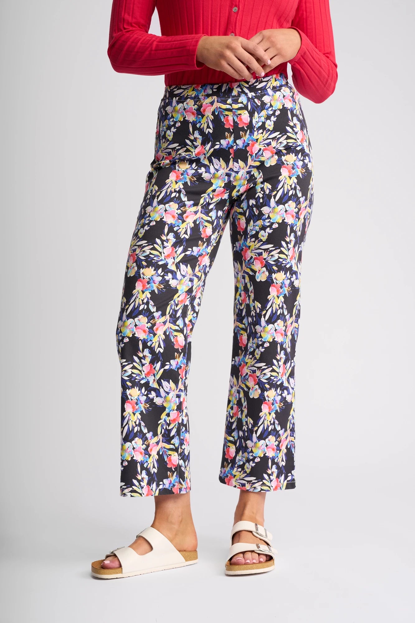 A person wearing a red long-sleeve top, white open-toe sandals, and black Vassalli Seattle Pull On - Lani pants crafted from stretch cotton with colorful floral prints stands against a plain background.