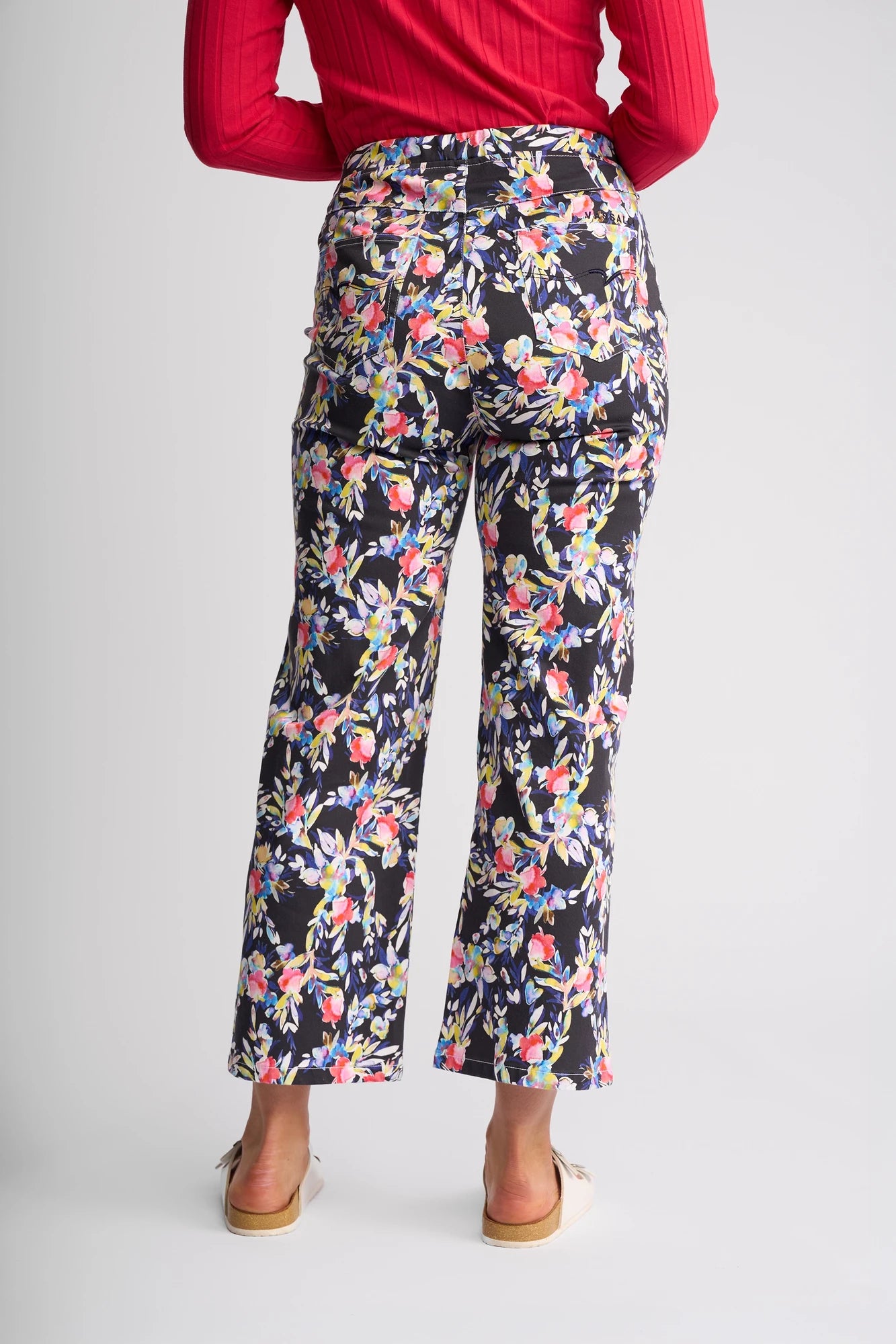 A person wearing the Vassalli Seattle Pull On - Lani black floral print pants made from comfortable stretch cotton, a red long-sleeve top, and beige sandals stands facing away from the camera against a plain white background.