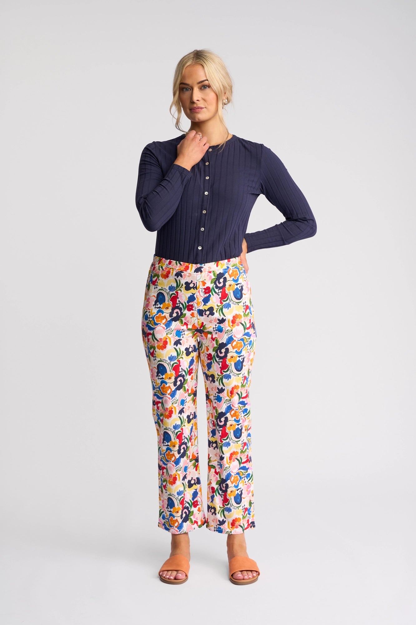 A woman stands against a plain background wearing a long-sleeve navy blouse, Vassalli Seattle Pull On - Mon Cheri floral print pants crafted from stretch cotton, and tan sandals. She has light hair tied back and rests one hand on her chin while the other is on her hip.