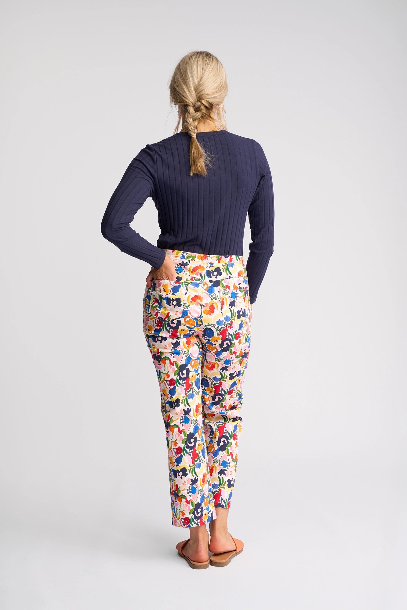 A woman with blonde hair in a braid, standing barefoot with her back to the camera against a plain light background, wears colorful Vassalli Seattle Pull On - Mon Cheri stretch cotton floral print pants and a navy long-sleeve top.