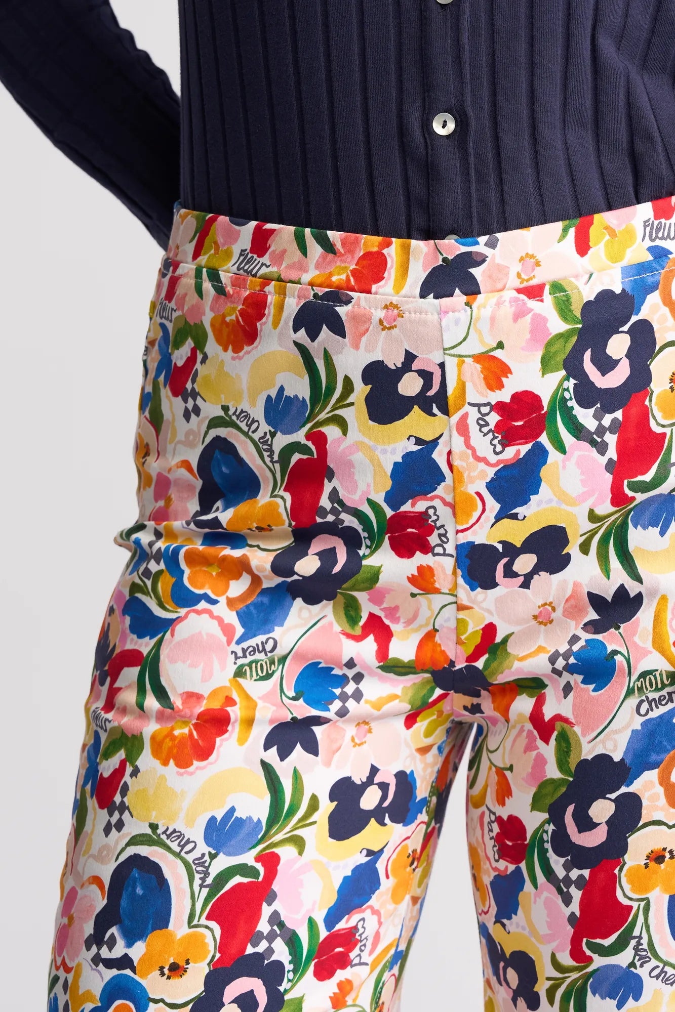 A person wears the Vassalli Seattle Pull On - Mon Cheri pants, featuring stretch cotton fabric adorned with vibrant floral prints and cursive writing on a white background, paired with a dark, long-sleeve ribbed shirt.