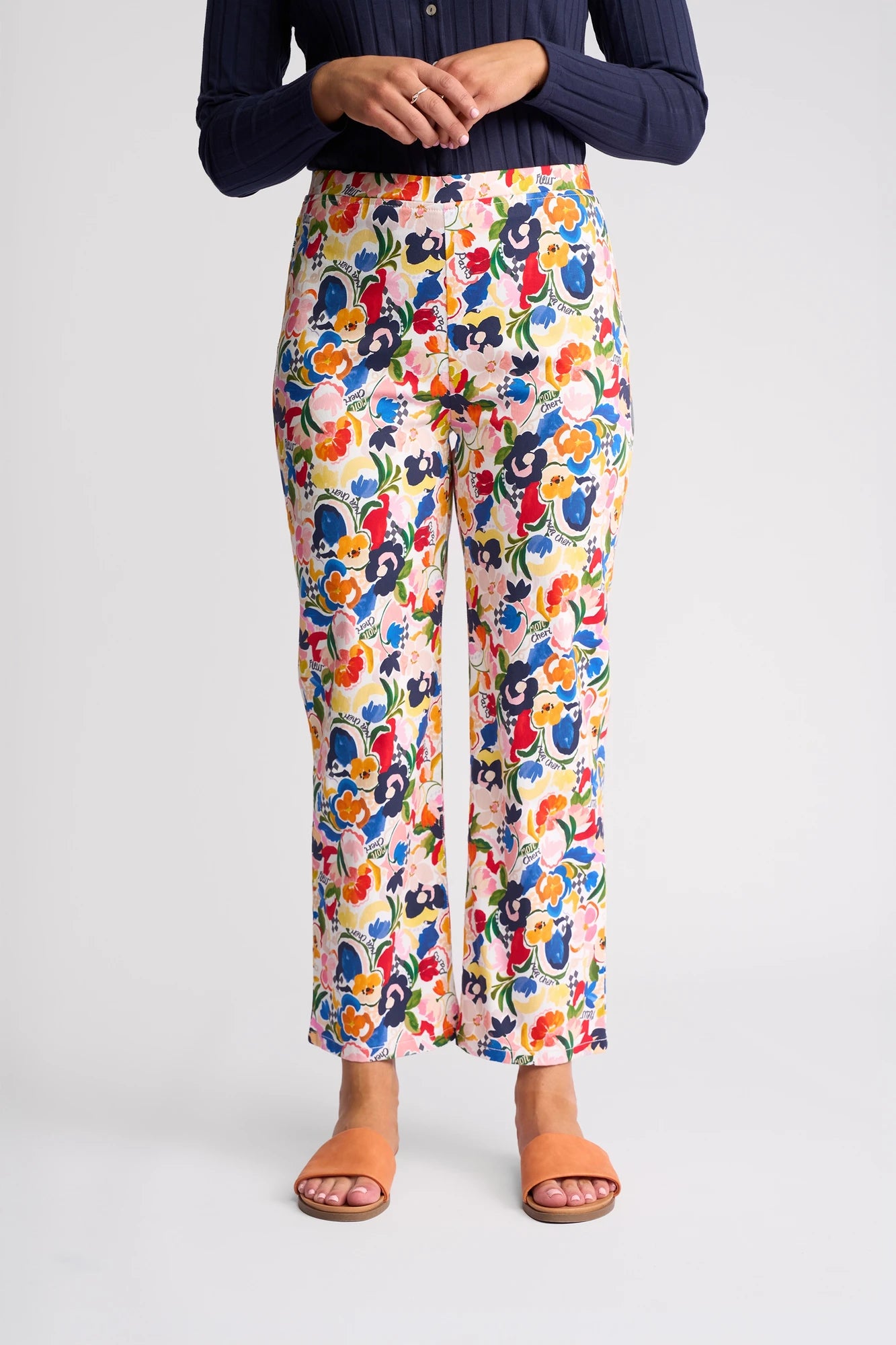 A person wearing vibrant Vassalli Seattle Pull On - Mon Cheri pants, featuring a floral print and made from stretch cotton, pairs them with a long-sleeve navy top and tan open-toe sandals while standing against a plain white background.