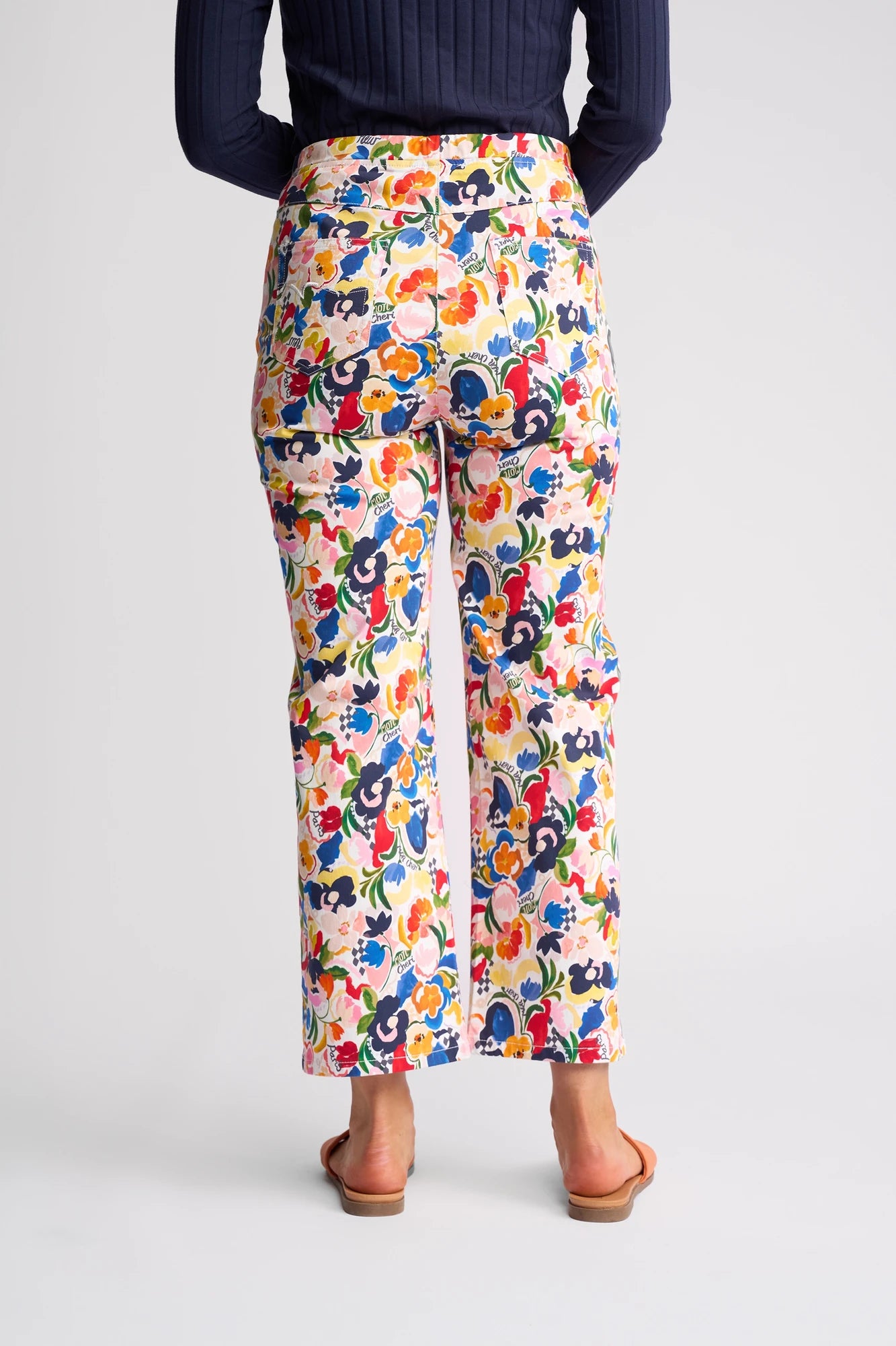 A person wearing the Vassalli Seattle Pull On - Mon Cheri high-waisted, wide-leg floral print pants, crafted from stretch cotton, paired with a dark long-sleeve top and sandals, is photographed from the back against a plain background.