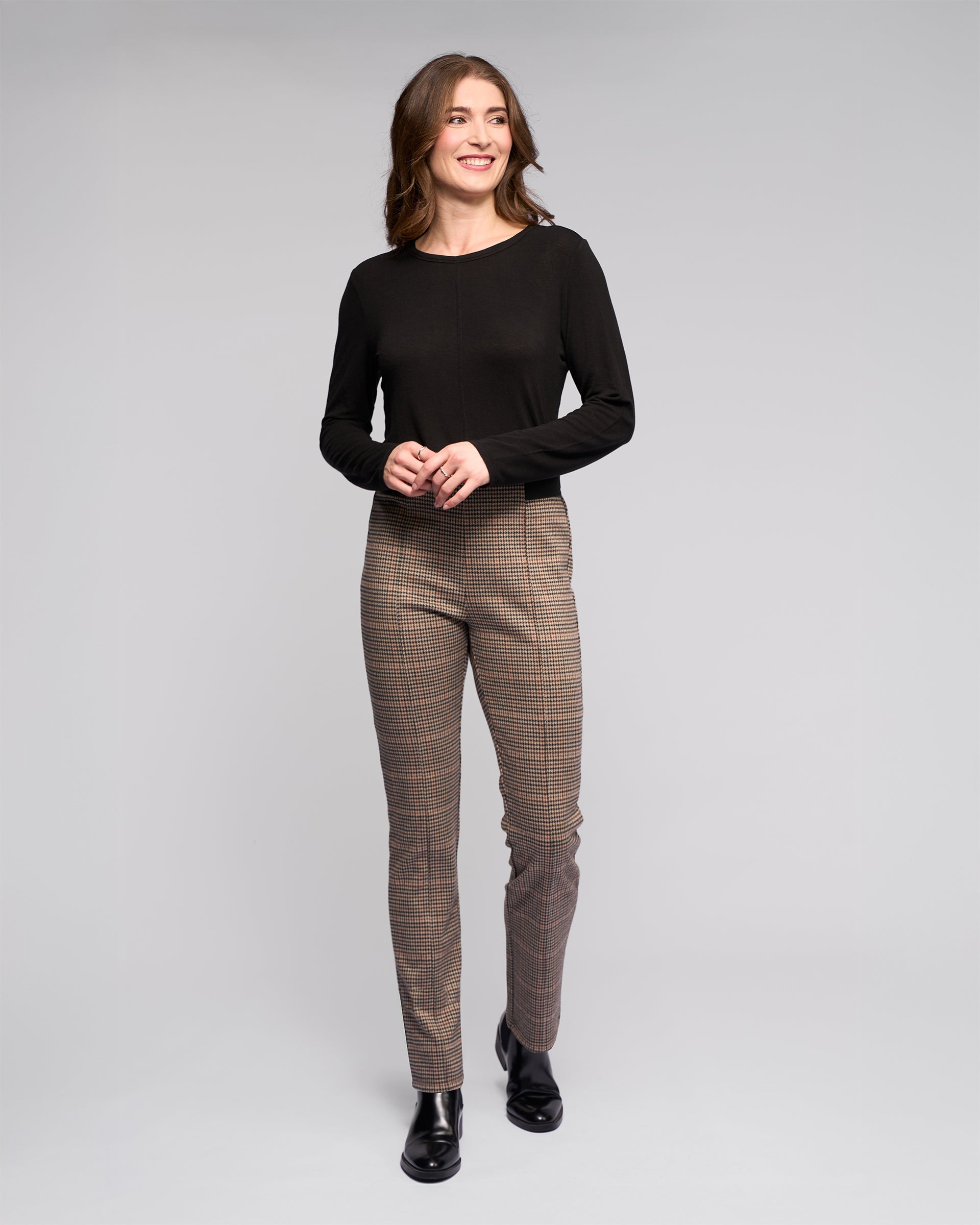 A woman smiles, looking to the side, wearing Vassalli's Everest Pull On - Ezra: a brown plaid slim pant in stretch knit fabric, paired with a black long-sleeve top and black boots, standing against a plain background with hands loosely clasped.
