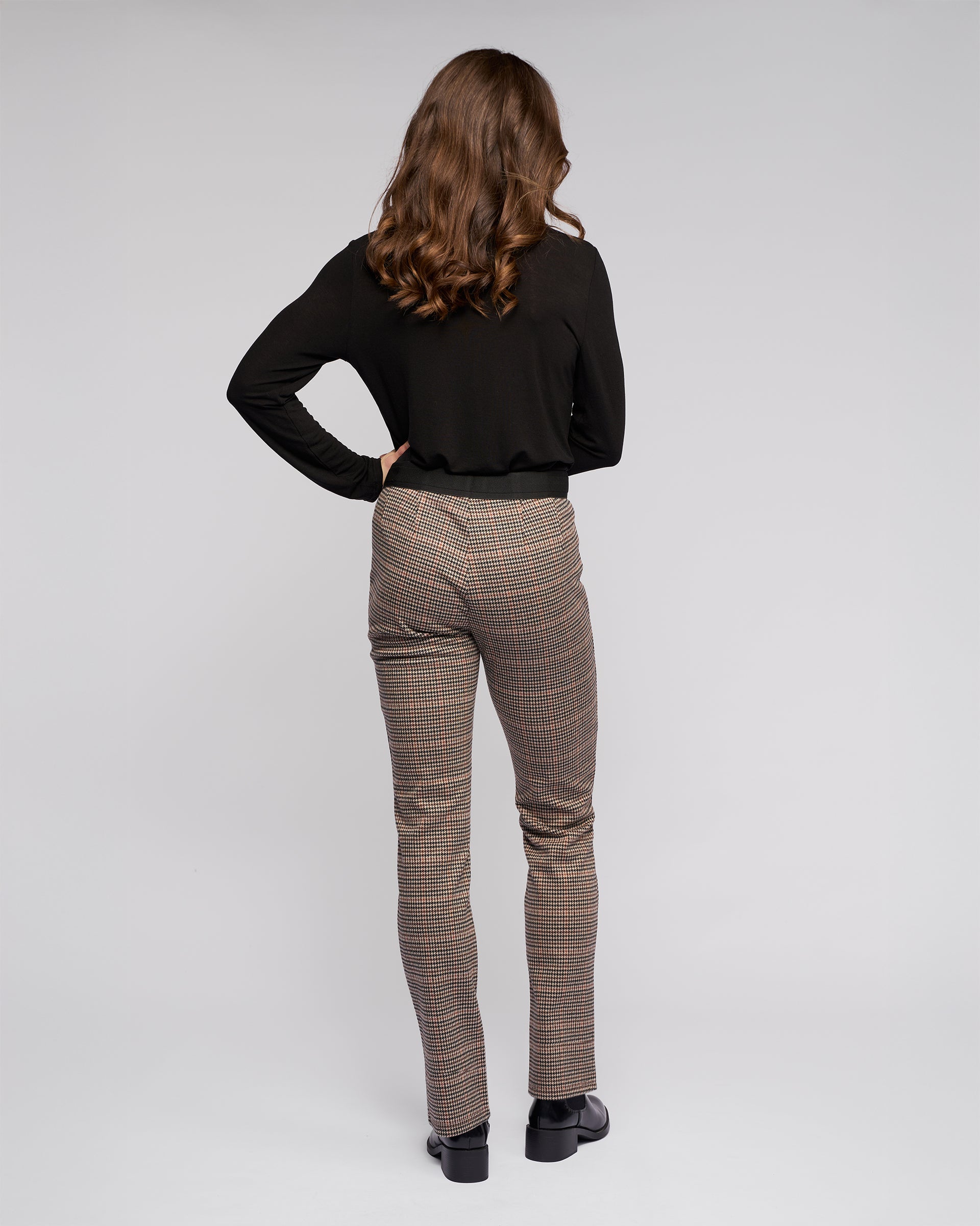A woman with wavy brown hair, facing away, wears a black long-sleeve top and Vassalli's Everest Pull On - Ezra print slim pants in stretch knit fabric with black shoes against a plain light gray background.