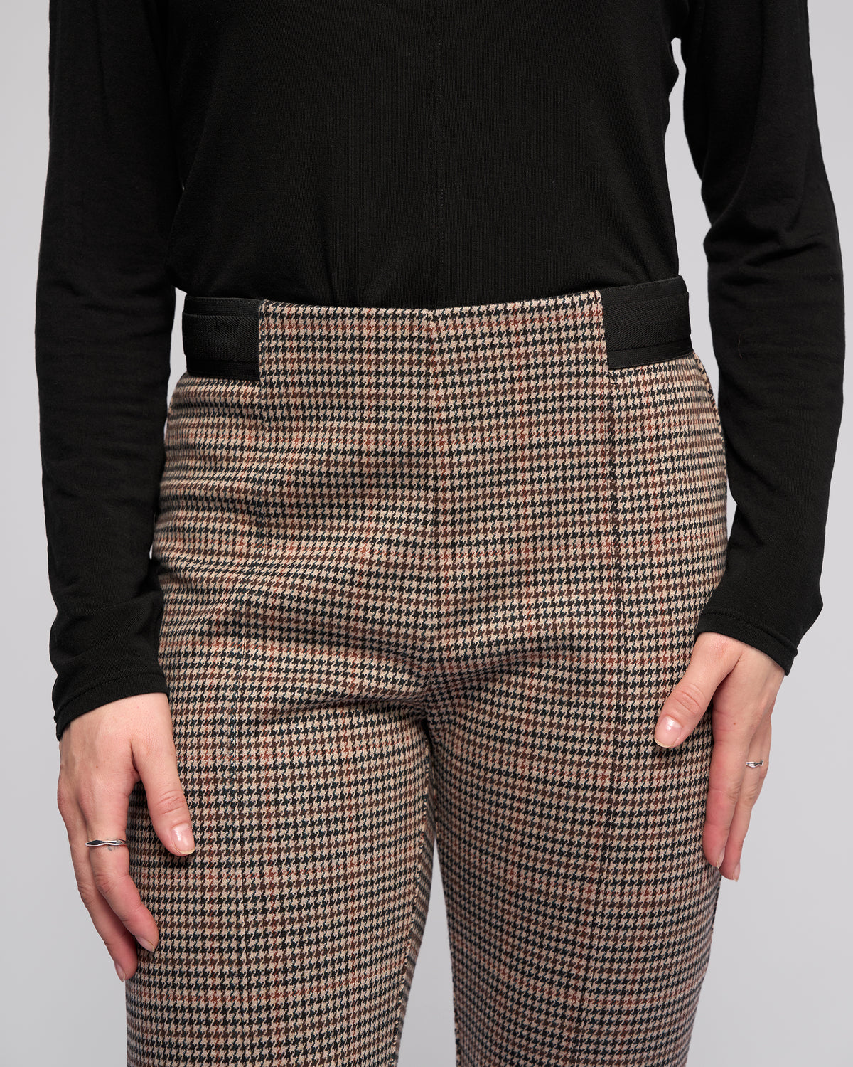 A person wearing the Vassalli Everest Pull On - Ezra slim pants, featuring a high waist and stretch knit fabric in a brown and black print, paired with a long-sleeved black top and hands resting on their thighs.