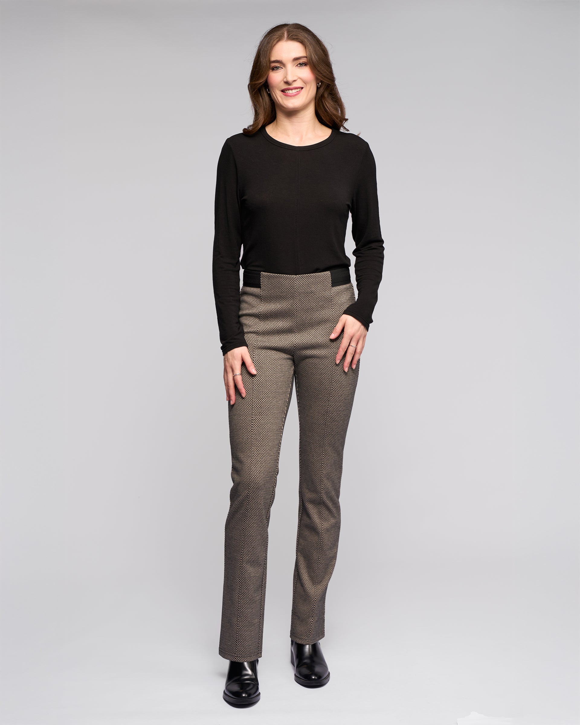 A woman with long brown hair smiles against a light gray background, wearing a black stretch knit top, black boots, and Vassalli Everest Pull On - Knox high-waisted slim pants.