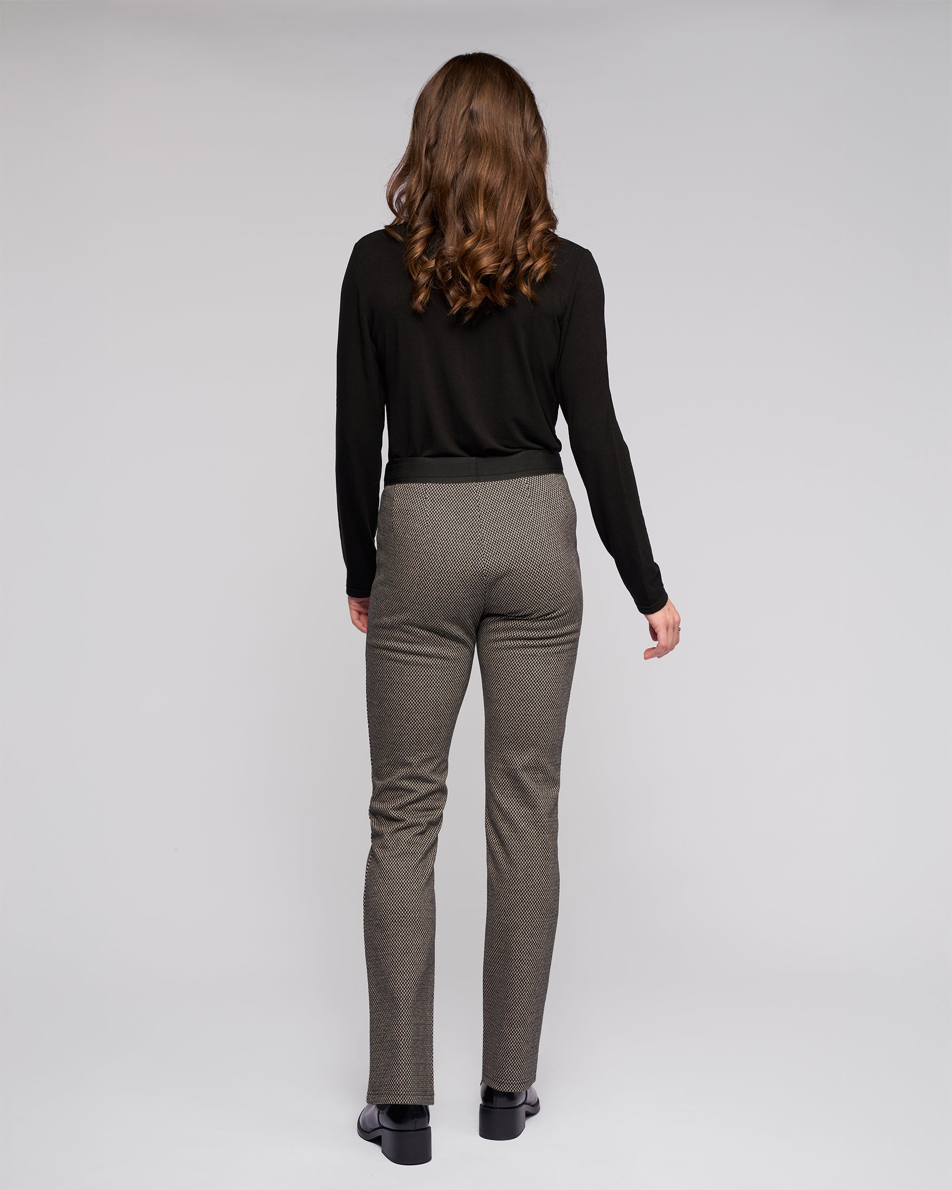 A woman with wavy brown hair faces away from the camera, wearing a long-sleeve black top, Vassalli Everest Pull On - Knox stretch knit gray slim pants, and black shoes, set against a plain light gray background.