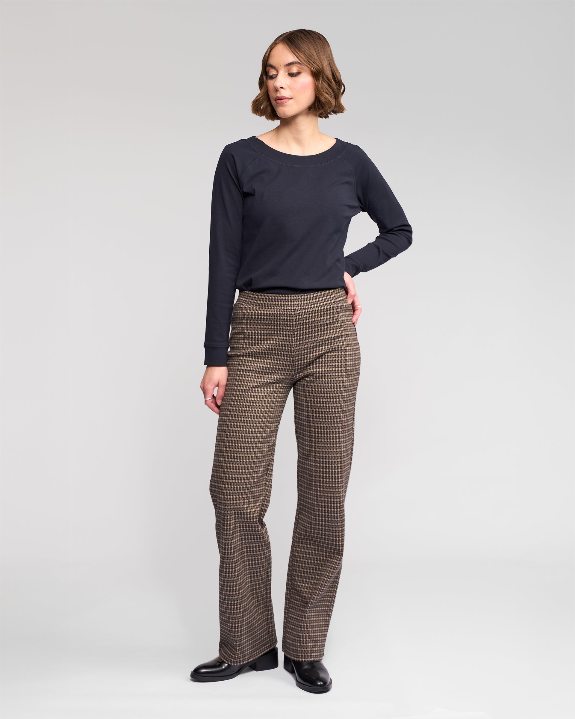 A woman with short brown hair poses with one hand on her hip against a light gray background. She wears a long-sleeve black top, Vassalli Byron Pull On - Nova pants, and black shoes, looking slightly to the side.