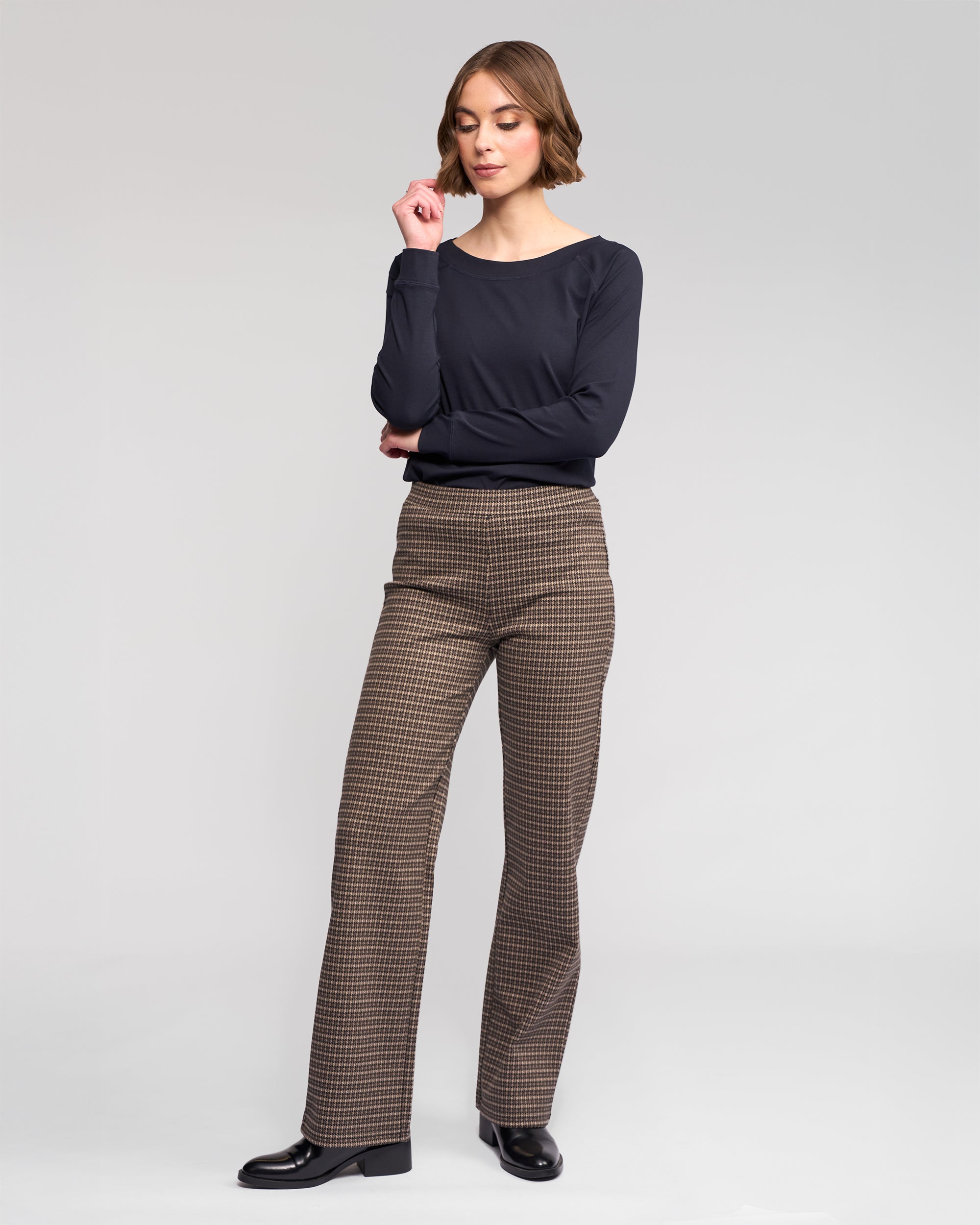 A woman with short, wavy hair stands in a neutral studio wearing Vassalli's Byron Pull On - Nova straight-leg pants, a long-sleeve black top, and black shoes. Her arms are relaxed, with one hand lifted near her face.