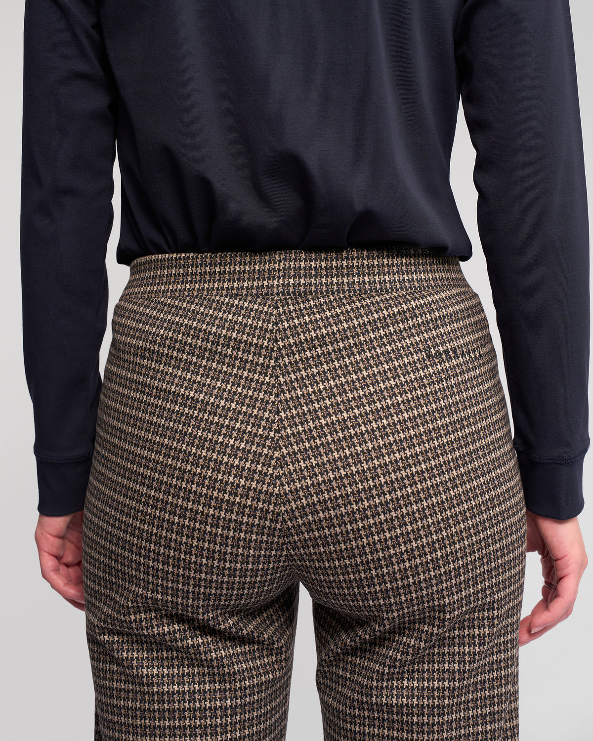 A person shown from the back, standing against a plain light background, wears a long-sleeve black top and Vassalli Byron Pull On - Nova straight-leg pants in brown houndstooth.