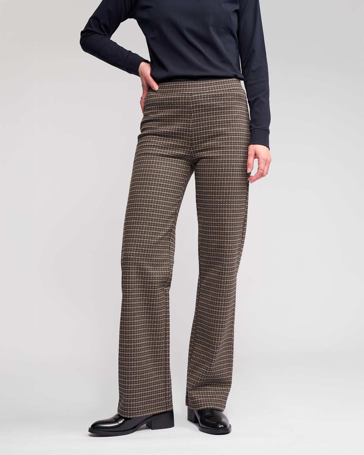 A person wearing Vassalli Byron Pull On - Nova stretch knit, houndstooth-patterned straight-leg pants, a long-sleeve black top, and black shoes stands with one hand on their hip against a plain light background.