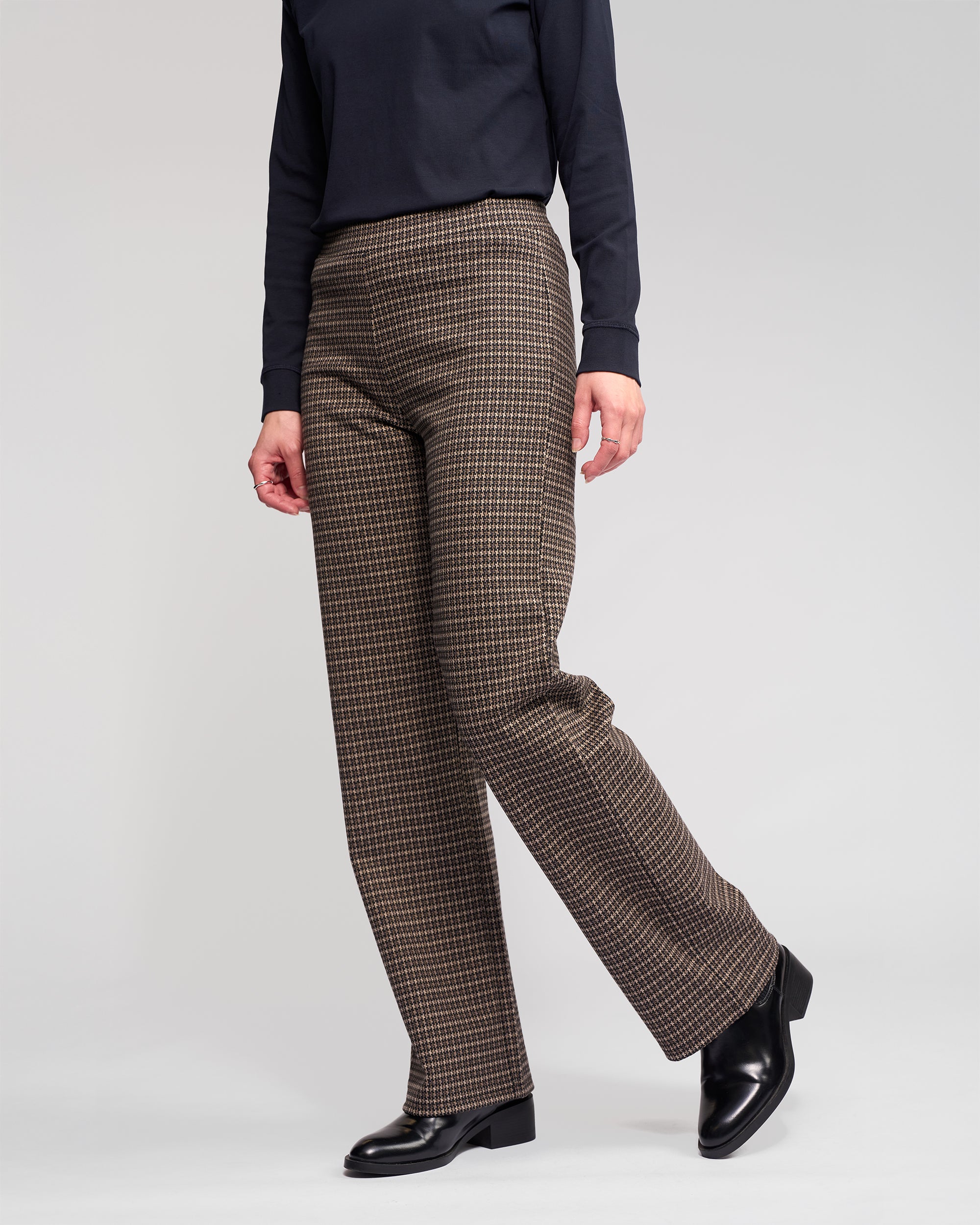 A person, face not visible, stands against a plain light background wearing a black long-sleeve top, Vassalli Byron Pull On - Nova straight-leg stretch knit pants in brown and black, and black heeled shoes.