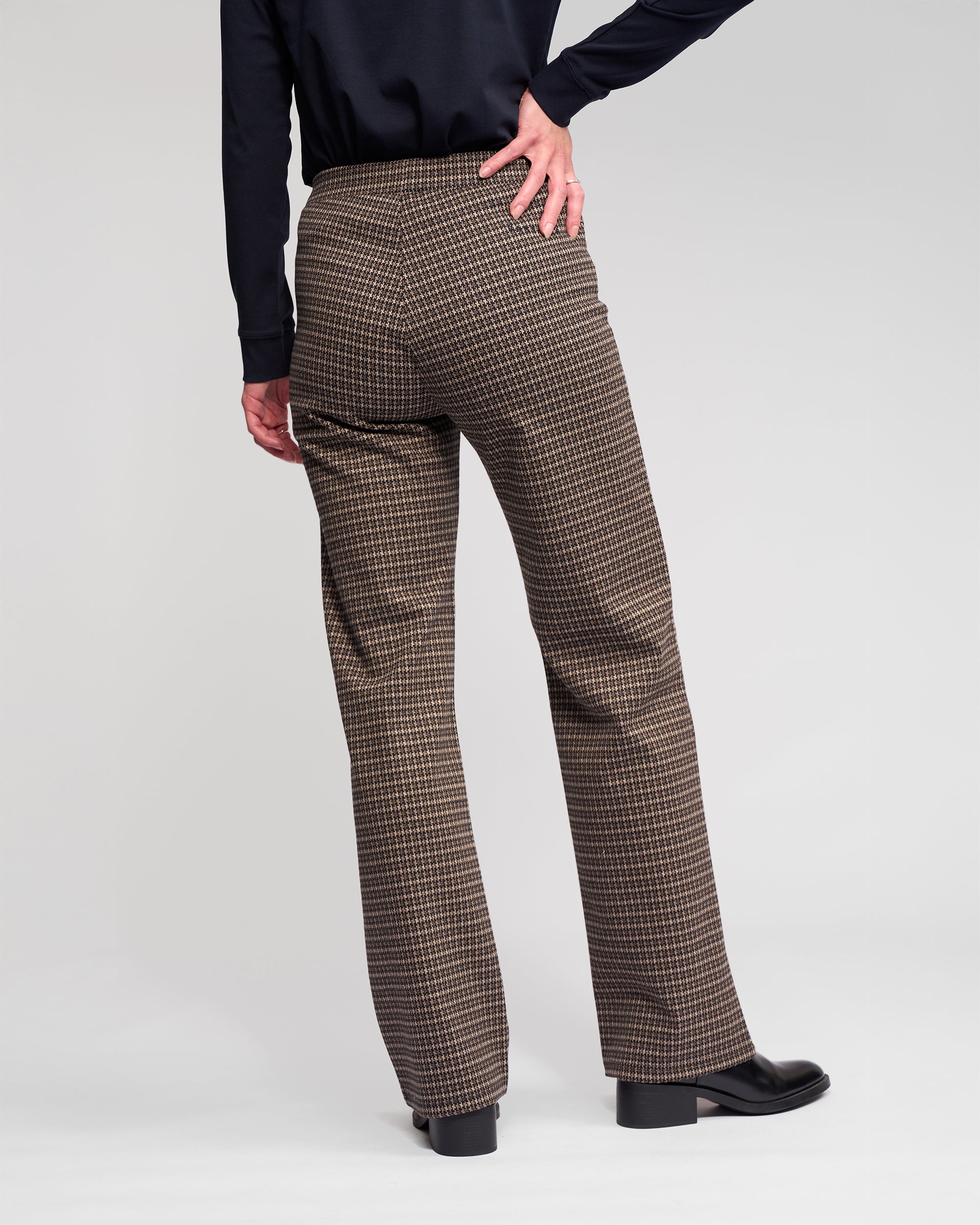 A person stands with one hand on their hip, wearing Vassalli Byron Pull On - Nova pants in a brown and black print, paired with a black top and shoes. The photo is taken from behind against a plain light background.