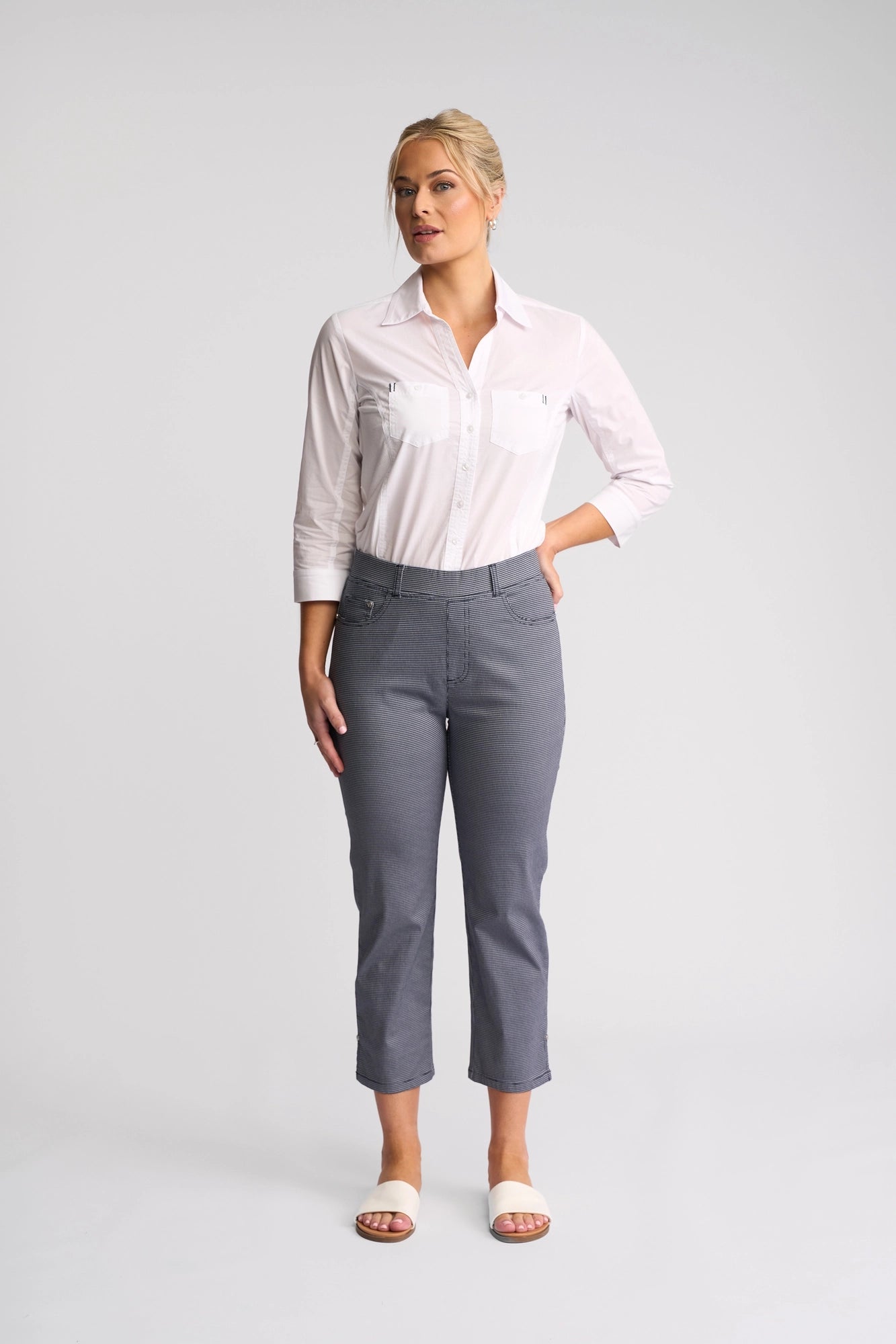 A woman stands against a plain background, wearing a white long-sleeve button-up shirt, Vassalli London Pull On pants in Midnight, and white open-toed shoes. She faces forward with one hand lightly touching her hip.