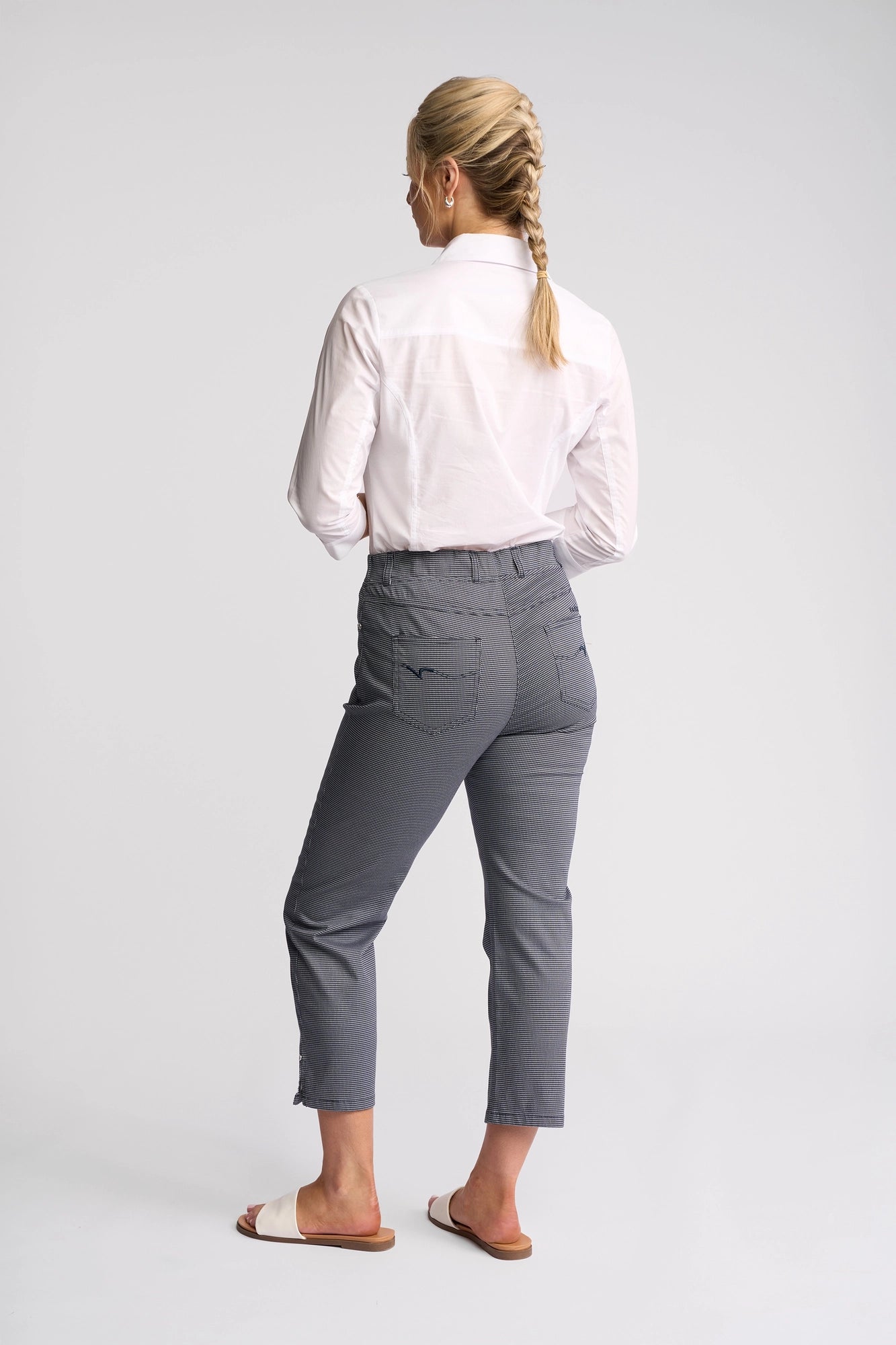 A person with blond hair in a braid wears a white long-sleeve shirt, Vassalli London Pull On - Midnight slim-leg pants, and white sandals, standing with their back to the camera against a plain light background.