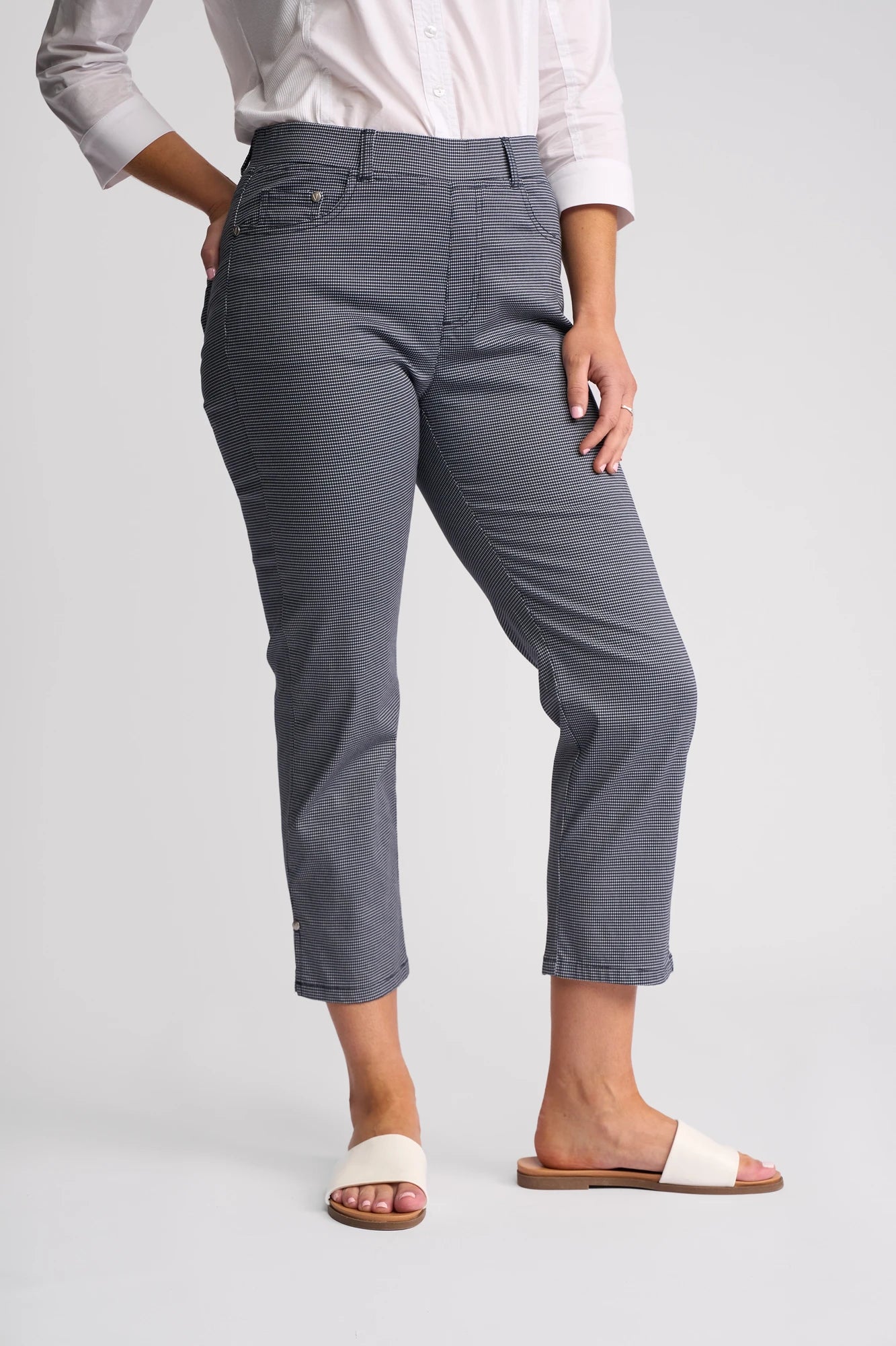 A person wearing Vassalli London Pull On slim-leg, ankle-length pants in Midnight, a white button-up shirt, and white slide sandals stands against a plain light background.