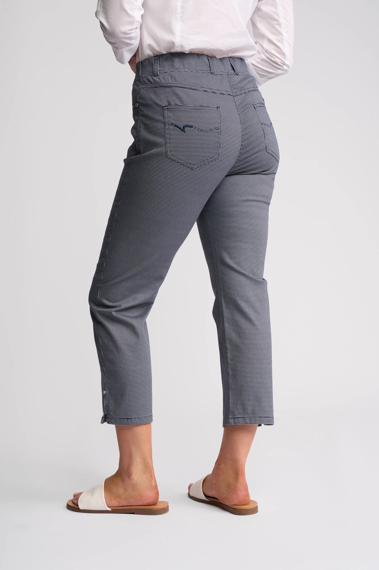 Wearing a crisp white shirt and Vassalli’s London Pull On - Midnight slim-leg pants with back pockets, the person stands barefoot except for beige slide sandals, facing away from the camera against a plain light background.
