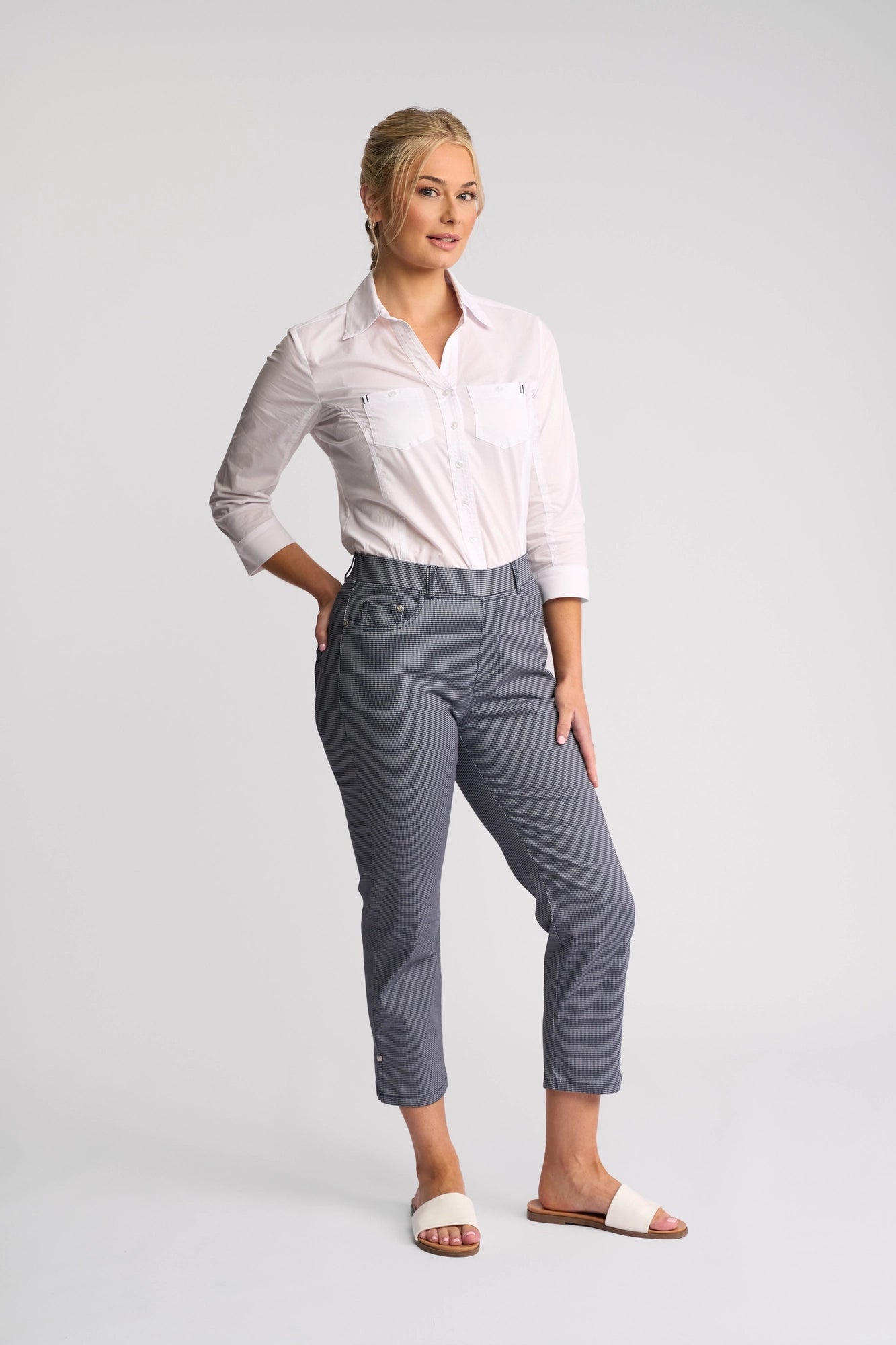 A woman stands against a plain background wearing a crisp white button-up shirt, Vassalli London Pull On Pants in Midnight, and white sandals. She has blonde hair styled up and is smiling, with one hand behind her back and the other resting on her thigh.