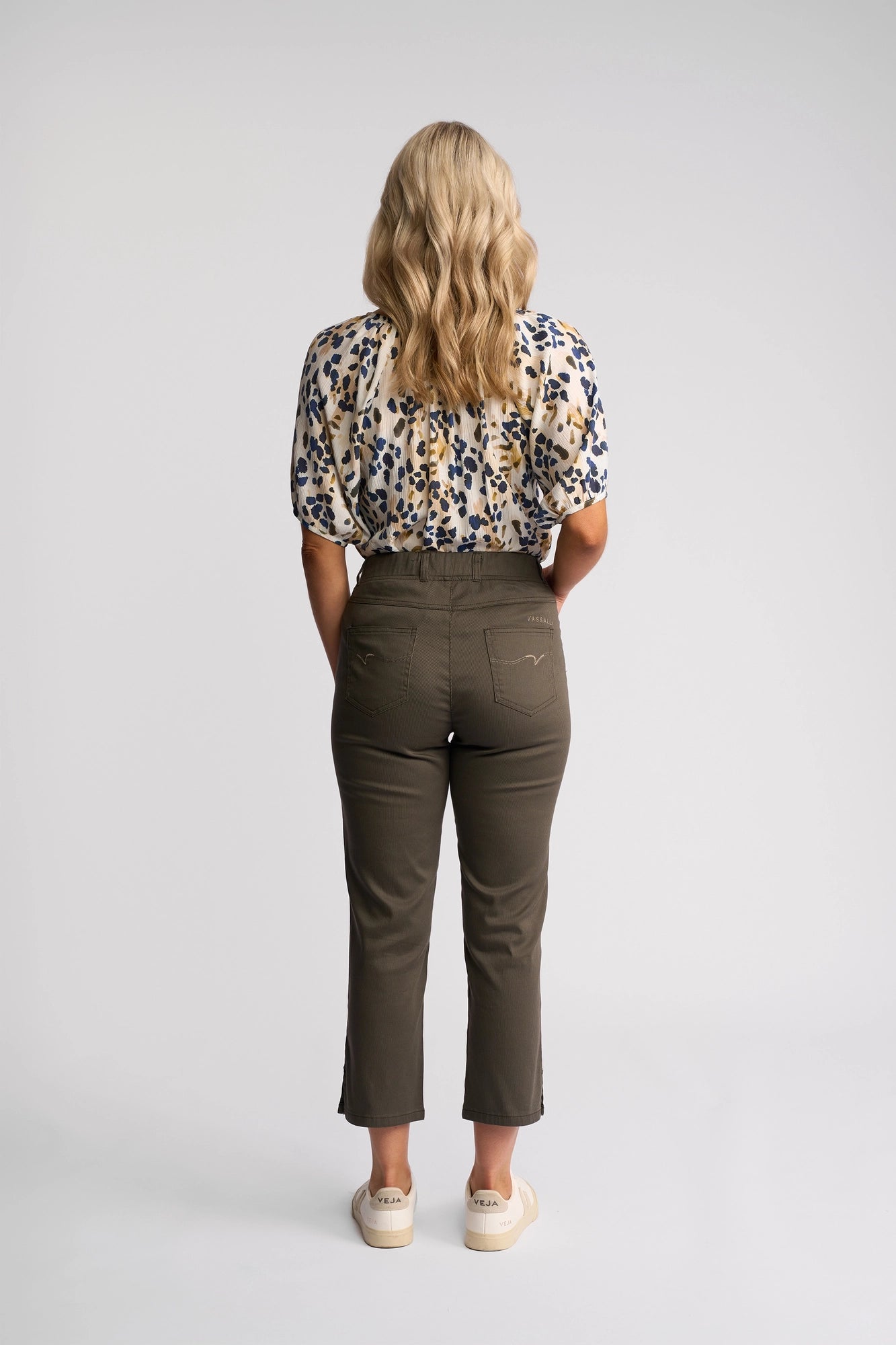 A person with wavy blonde hair stands facing away, wearing a floral top, white shoes, and the Vassalli London Pull On - Moss slim-leg pants in lightweight cotton, set against a plain light background.