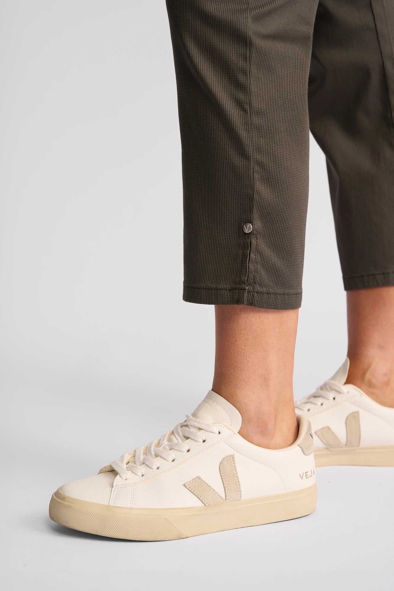A person wearing the Vassalli London Pull On pants in moss, paired with cream-colored Veja sneakers featuring beige soles, is standing on a light surface. Only their lower legs and feet are visible.