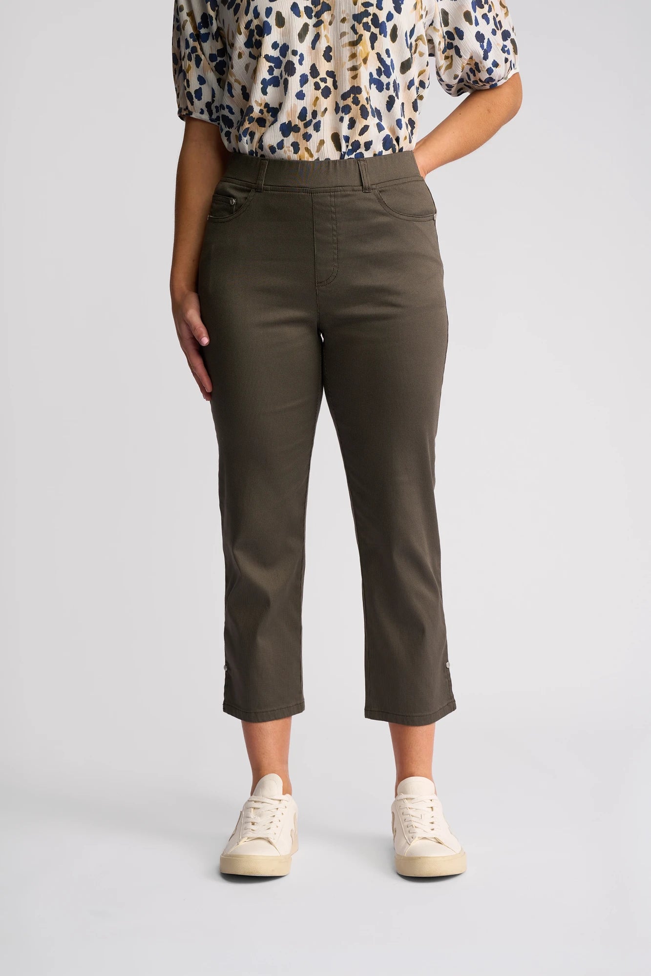 A person wearing an animal print blouse, Vassalli’s London Pull On - Moss slim-leg cropped pants in a lightweight cotton style, and cream sneakers stands against a plain light background, facing forward with hands in pockets.