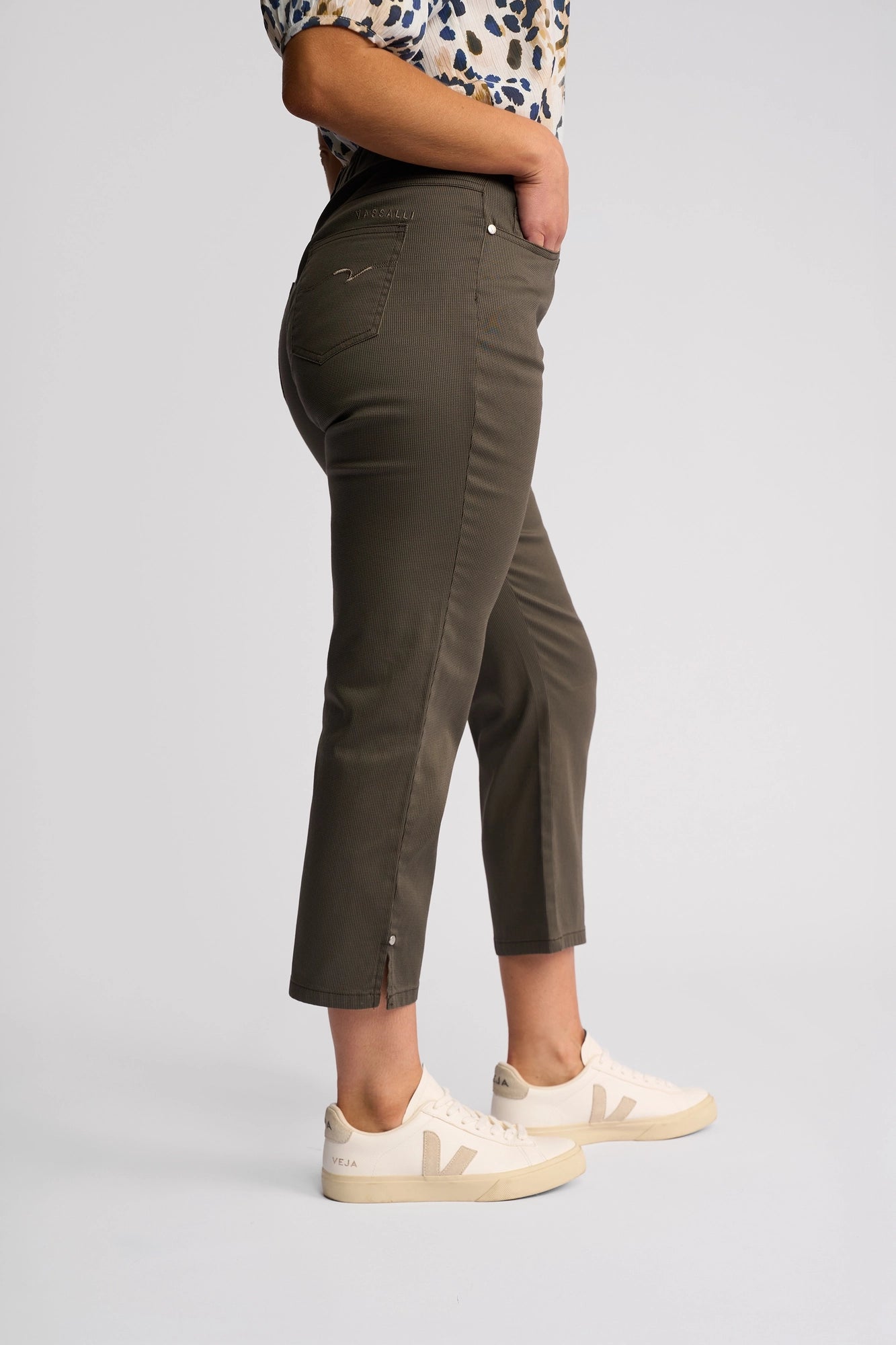 Wearing the Vassalli London Pull On pants in Moss, a person stands sideways against a plain background, pairing the olive green slim-leg cropped trousers with a patterned short-sleeve shirt and white sneakers.