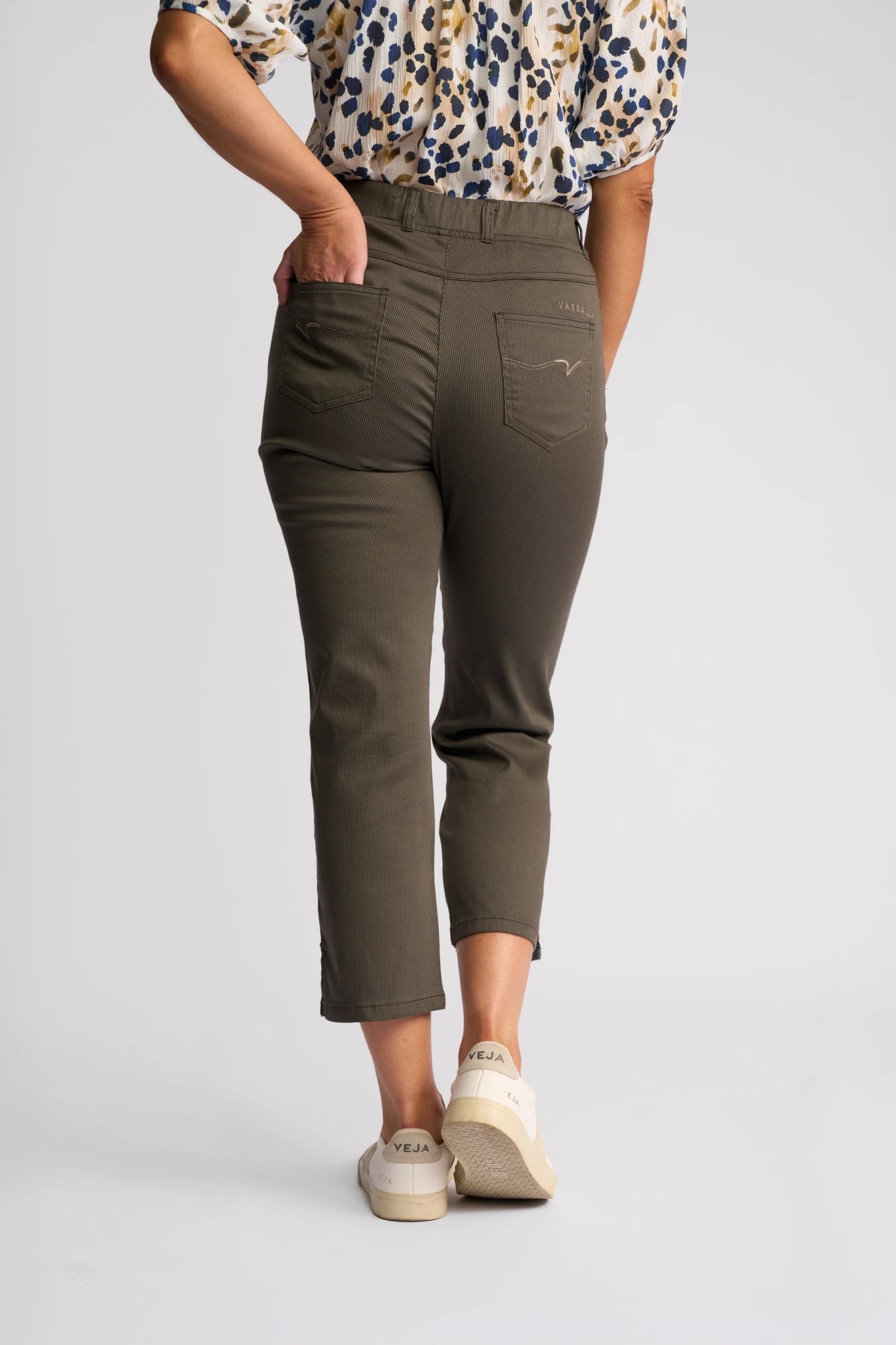 Wearing the Vassalli London Pull On - Moss pants, a person stands with one hand in their back pocket, facing away from the camera. The olive green cropped slim-leg style is paired with a patterned blouse and white Veja sneakers against a plain background.
