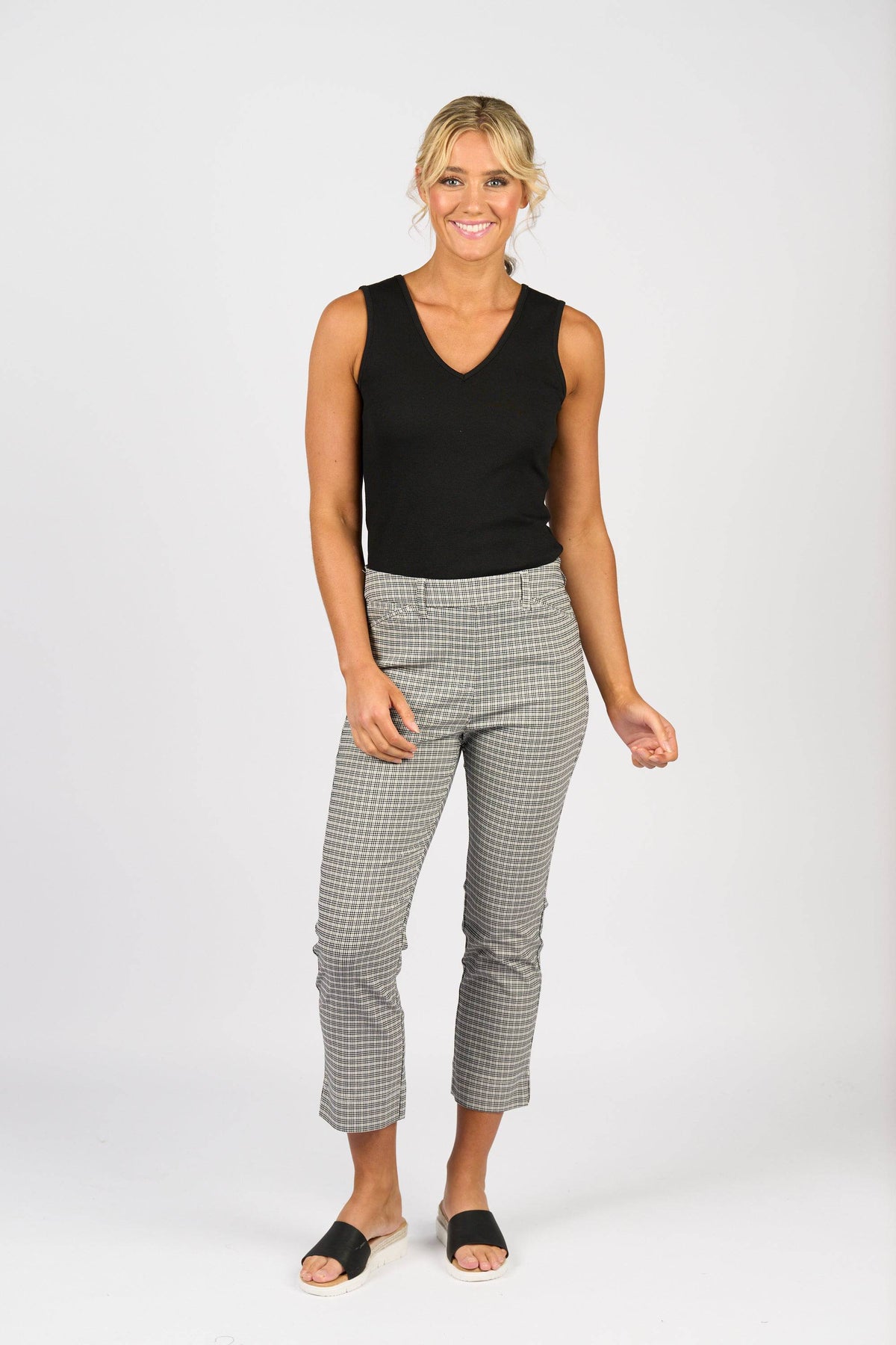 A woman smiles against a plain white background, wearing Vassalli’s Tint Pull On - Mini Check slim leg pants with a sleeveless black top and black slide sandals. Her blonde hair is tied back, radiating relaxed confidence.