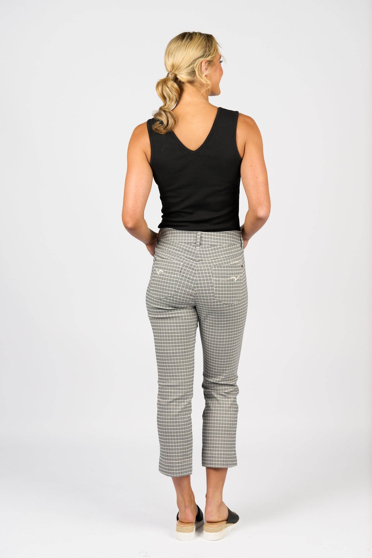 A woman with blonde hair in a low ponytail stands facing away, wearing Vassalli's Tint Pull On - Mini Check slim leg pants, a black sleeveless top, and black slip-on shoes against a plain white background.