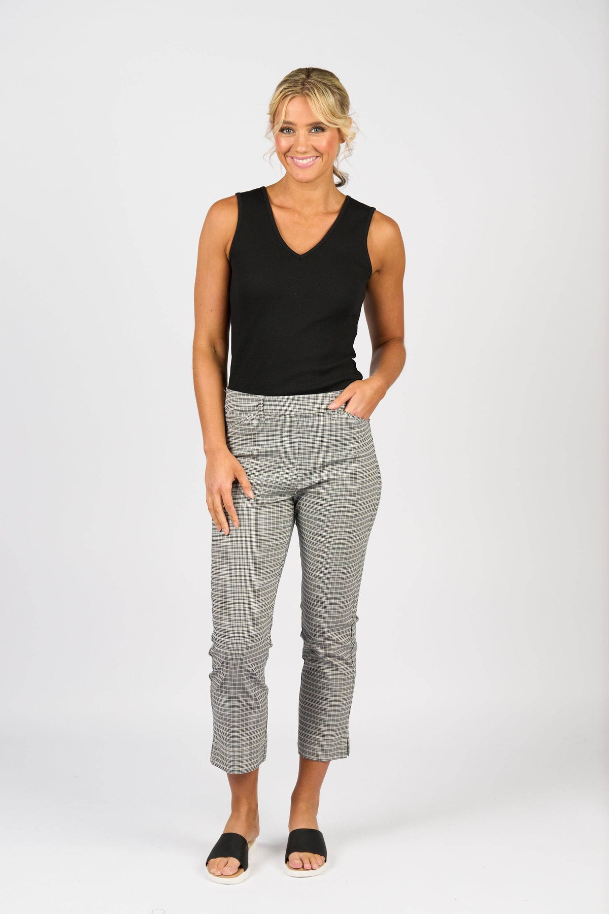 A woman smiles against a white background, wearing Vassalli Tint Pull On - Mini Check pants with a sleeveless black top and black open-toe shoes. One hand is in her pocket while the other rests by her side.