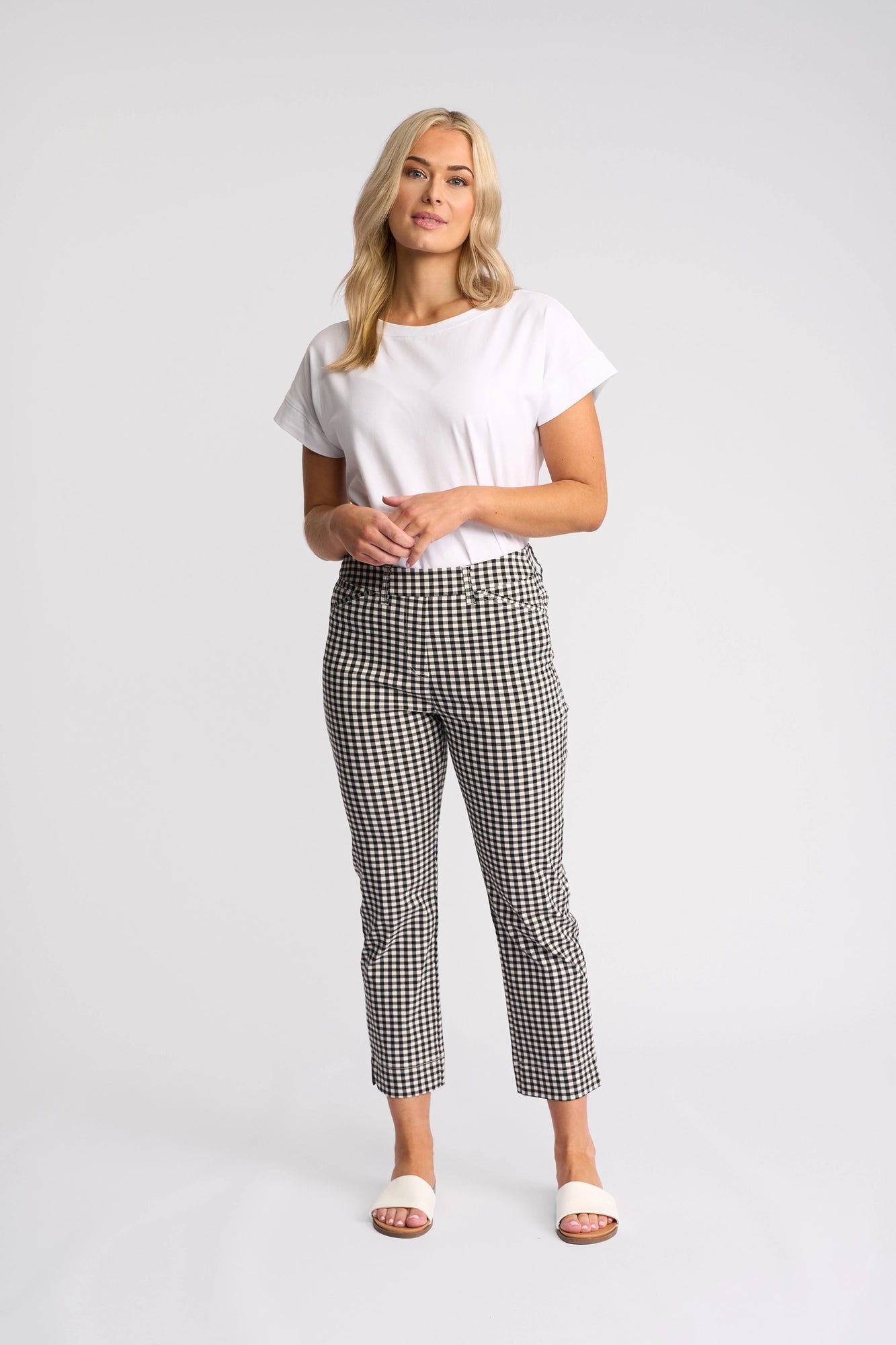 A woman stands against a plain background, wearing a white t-shirt paired with the Vassalli Tint Pull On pants in black gingham, featuring a slim leg design and pull-on style. She finishes the look with white sandals. Her blonde hair is styled down as she looks forward with a neutral expression.