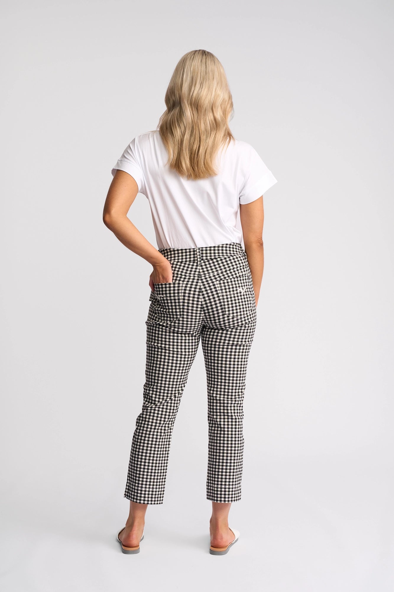 A person with long blonde hair, wearing a white t-shirt, Vassalli Tint Pull On - Black Gingham pants, and white slip-on shoes, stands facing away with one hand on their hip against a plain background.