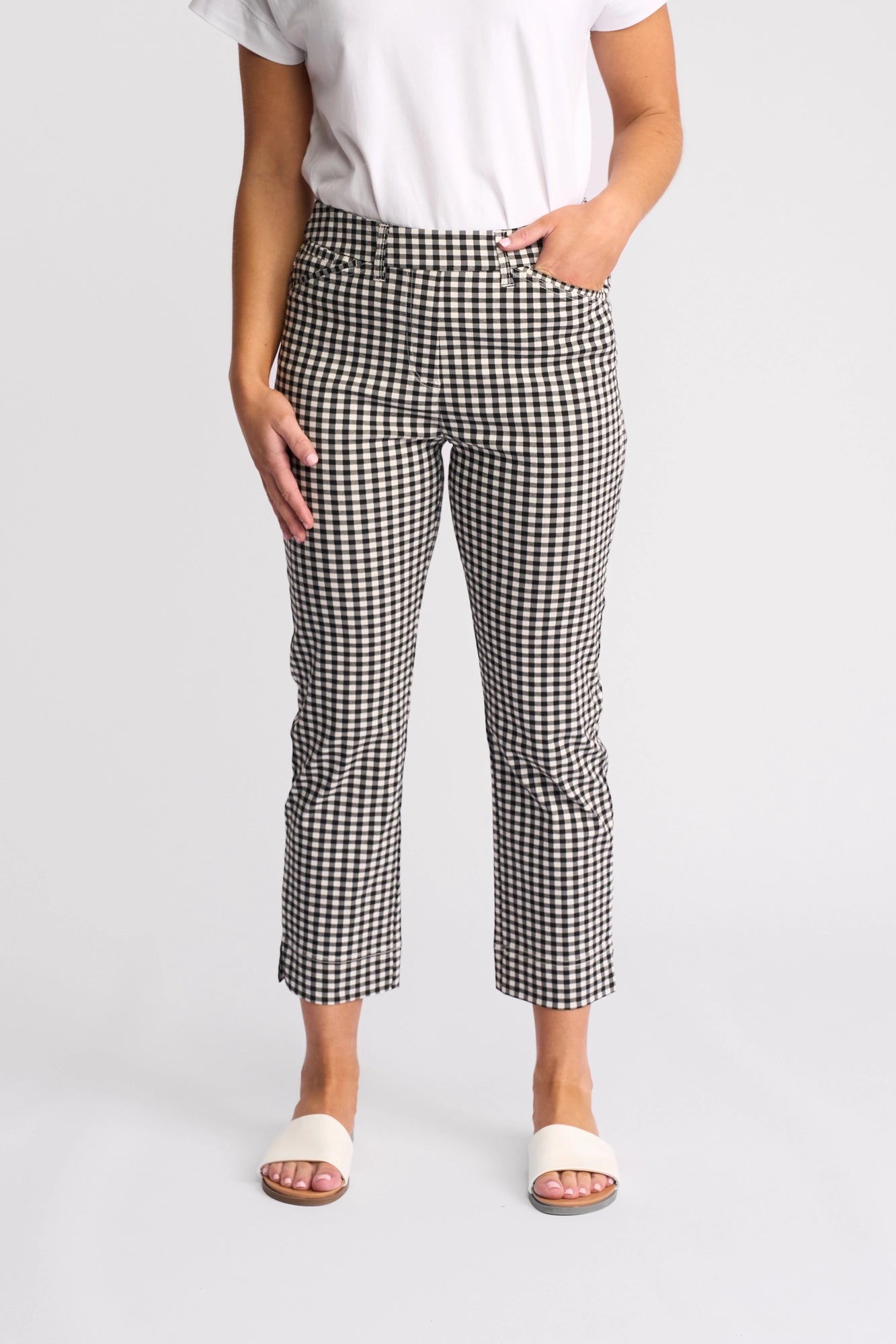 A person stands against a plain background with one hand in their pocket, wearing a white short-sleeve top, Vassalli’s Tint Pull On pants in Black Gingham, and white open-toe sandals.