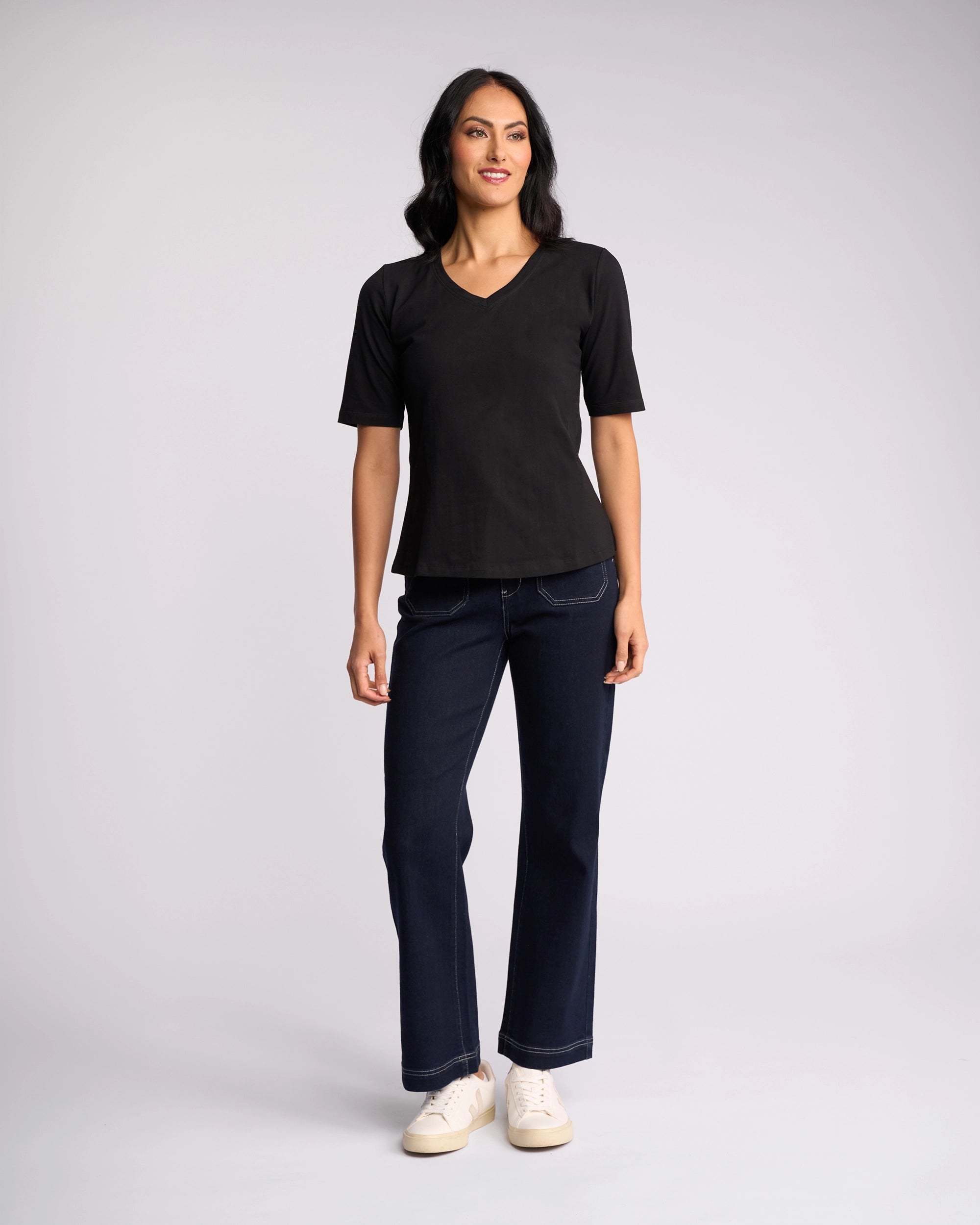 A woman stands against a plain background, smiling with arms at her sides, wearing a Vassalli Base Tee - Black with a V-neckline, paired with dark blue jeans and white sneakers.