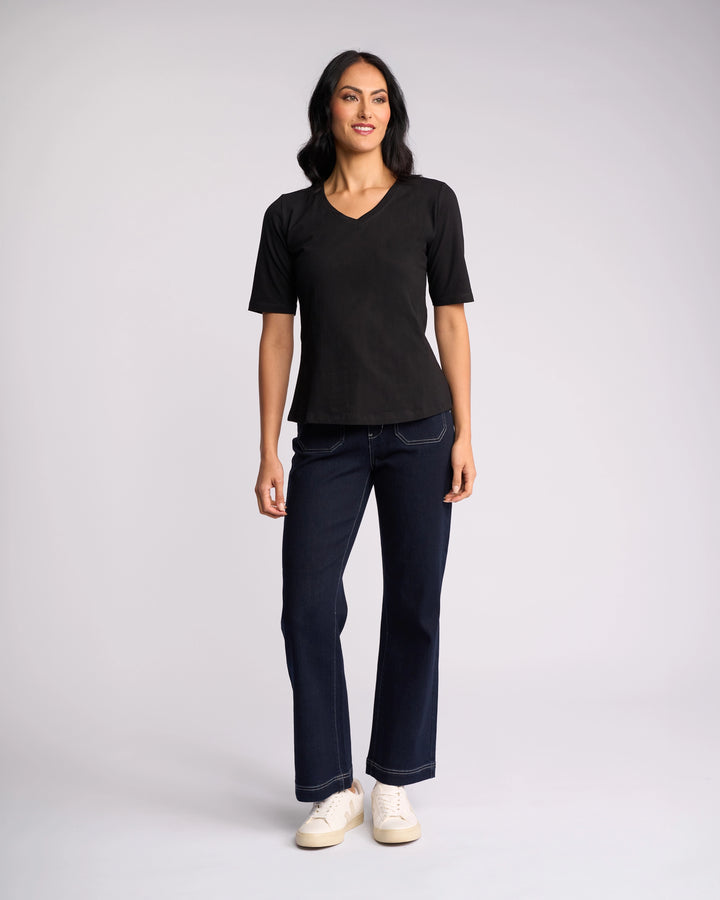 A woman stands against a plain background, smiling with arms at her sides, wearing a Vassalli Base Tee - Black with a V-neckline, paired with dark blue jeans and white sneakers.