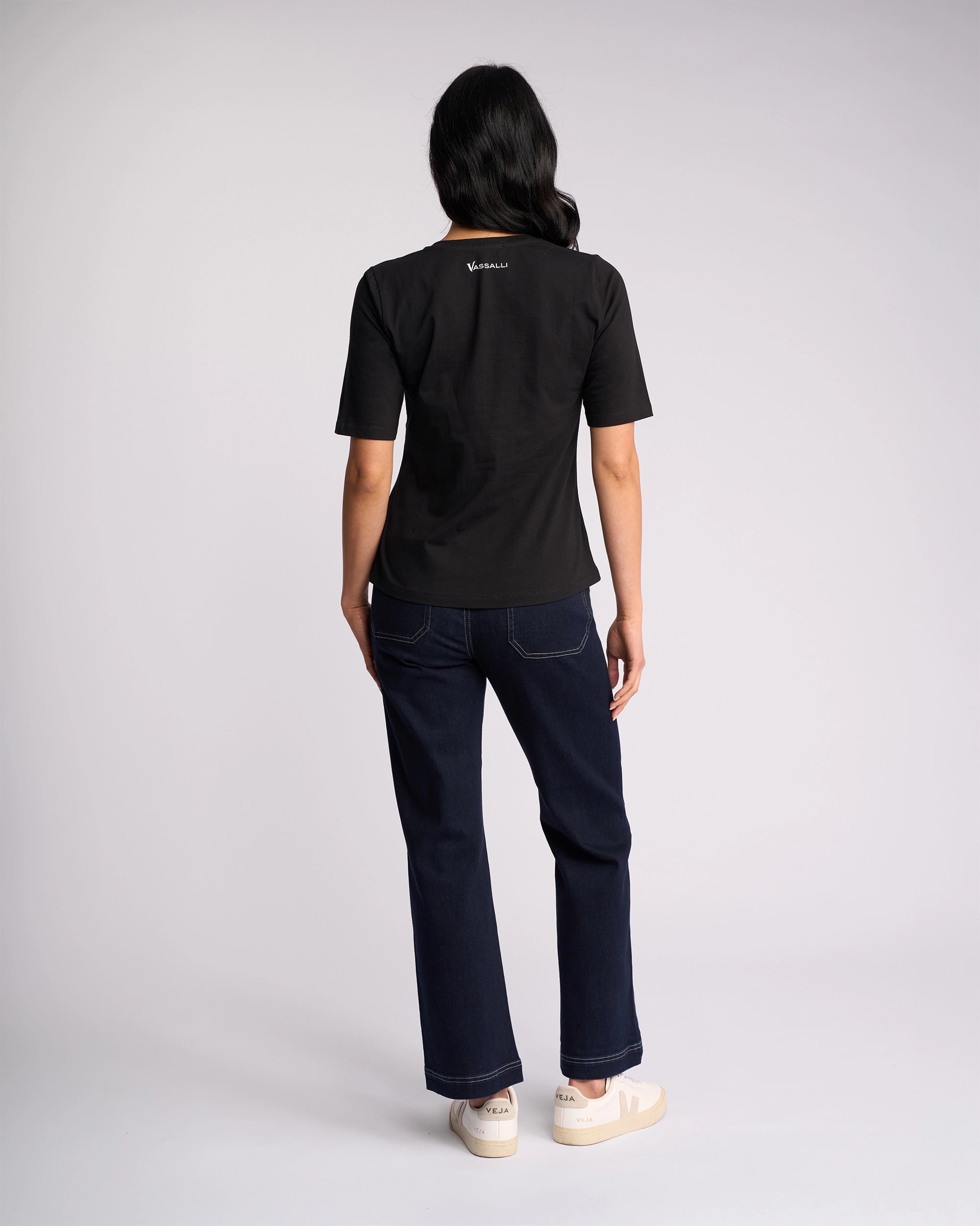 A person with long dark hair faces away, wearing the Vassalli Base Tee - Black, paired with dark blue jeans and white sneakers, set against a plain light background.